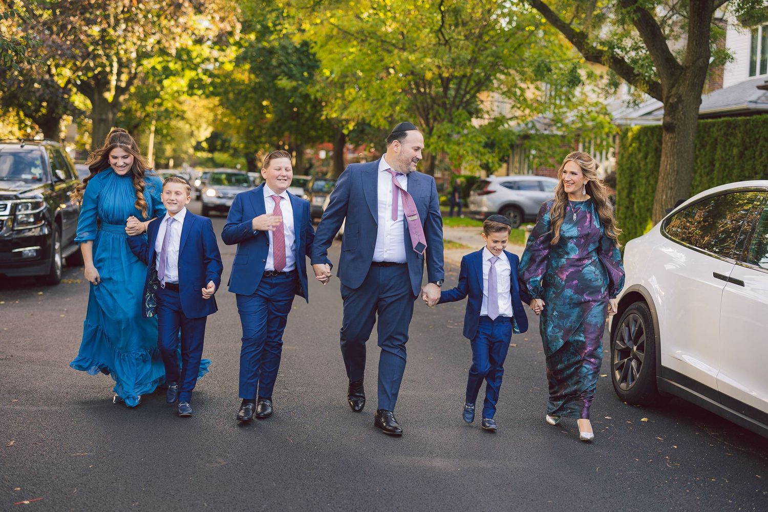 A family group walks together outside on a tree-lined street, dressed in formal attire