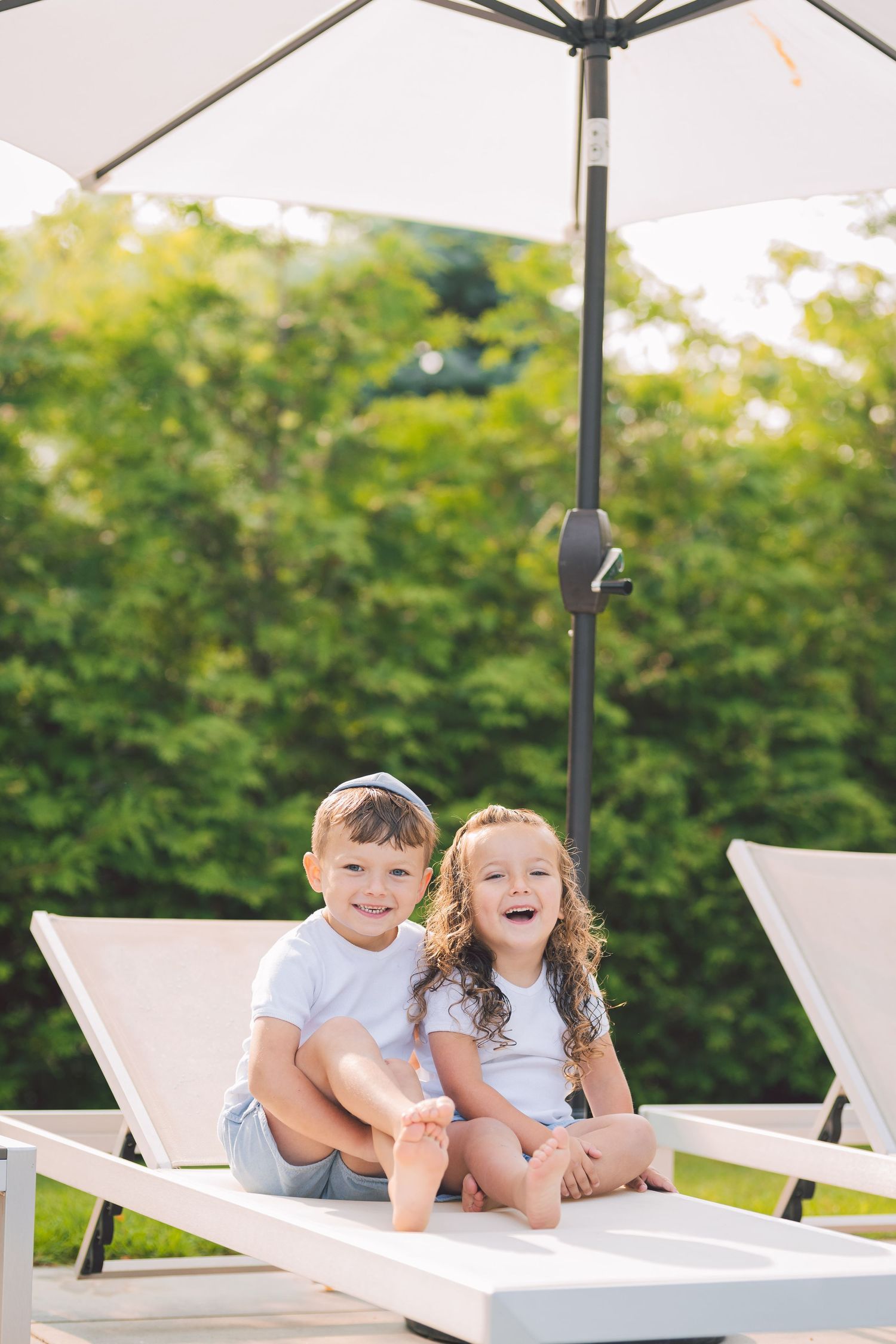 Children laughing while relaxing on white lounge chairs under an umbrella.