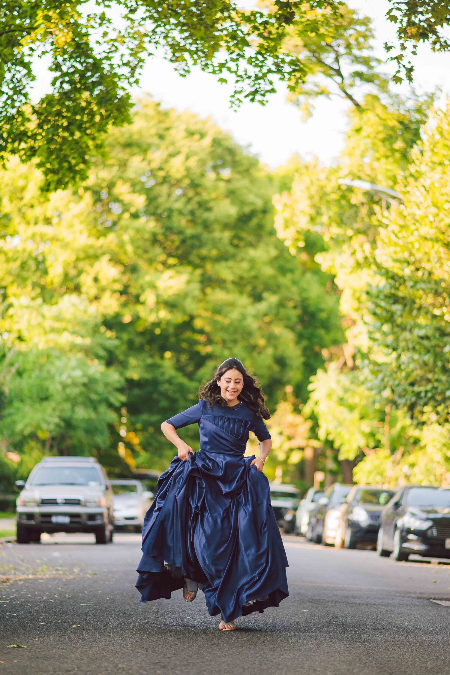 Person in navy blue formal dress spinning dramatically on tree-lined street at sunset.