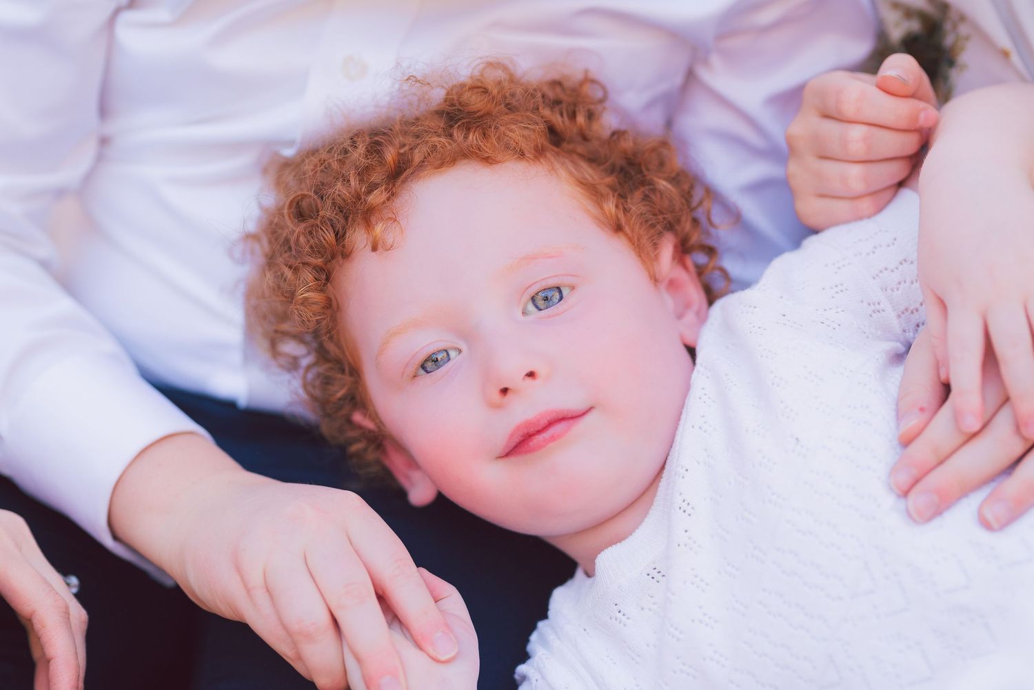 Close-up portrait of a young child with curly red hair lying on white bedding.