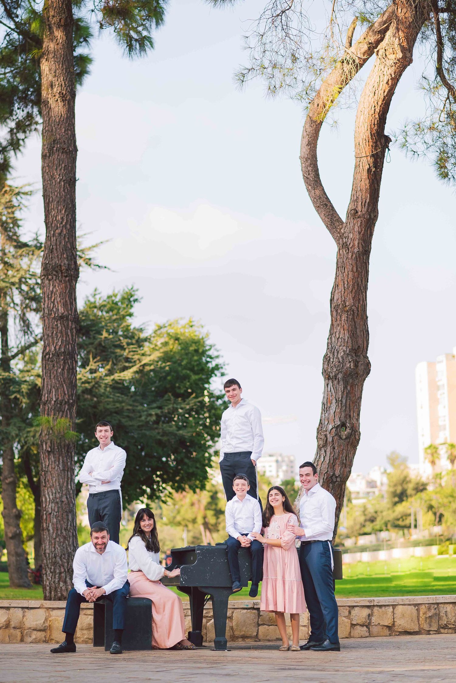 Group of people gathered casually in a park with pine trees and stone walls at sunset.