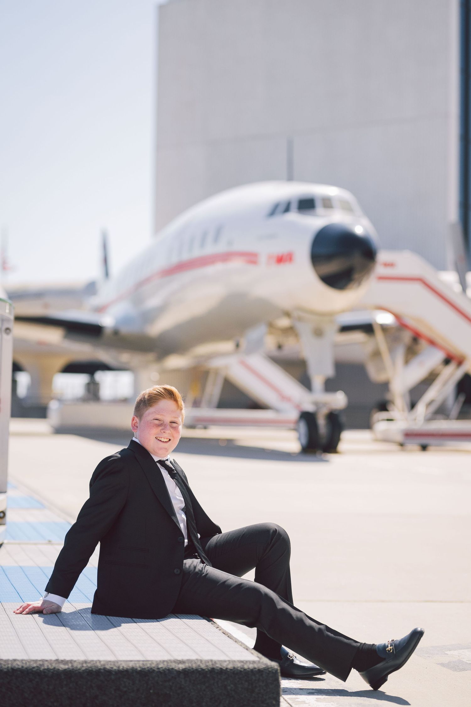 Person in formal attire sitting on concrete with vintage aircraft in background.