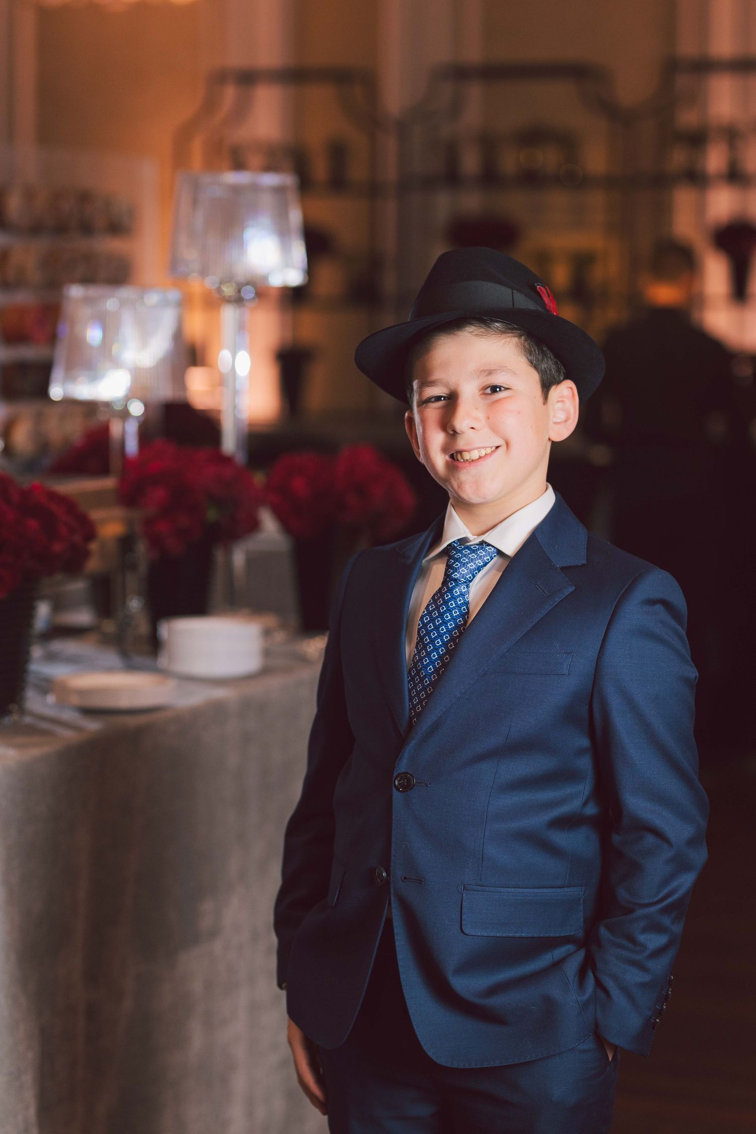 Young person in navy blue suit and polka dot tie poses elegantly at formal event with red floral decor.