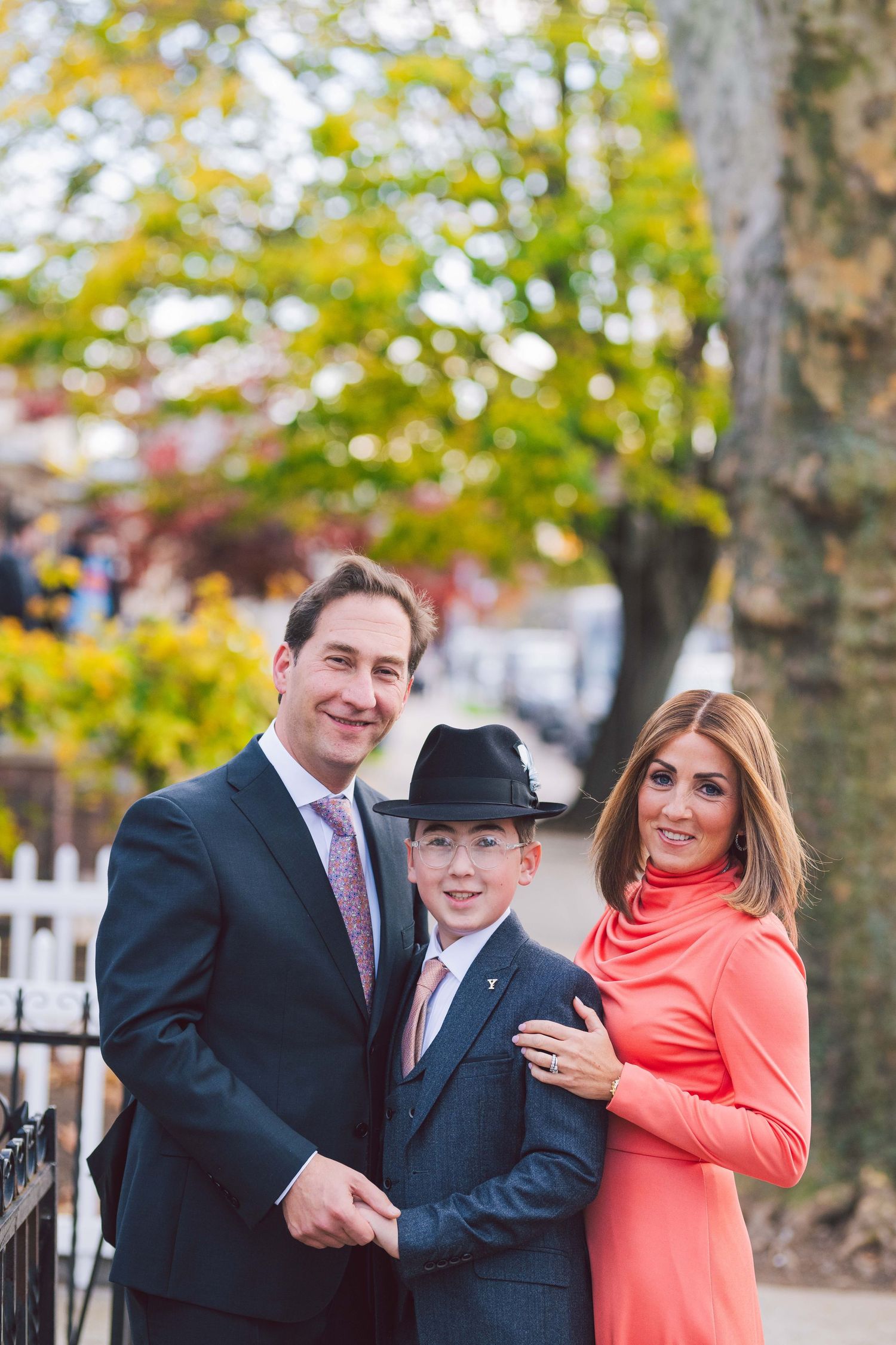 Family portrait taken outdoors during autumn with trees and iron fence in background.