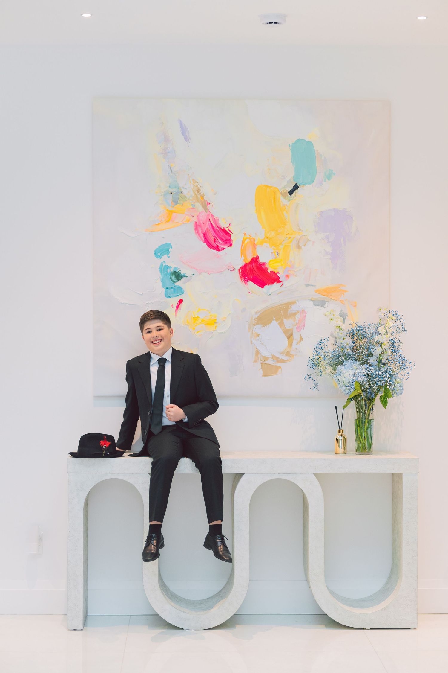 Young person in black suit sitting on modern white console table before colorful abstract painting with blue flowers.