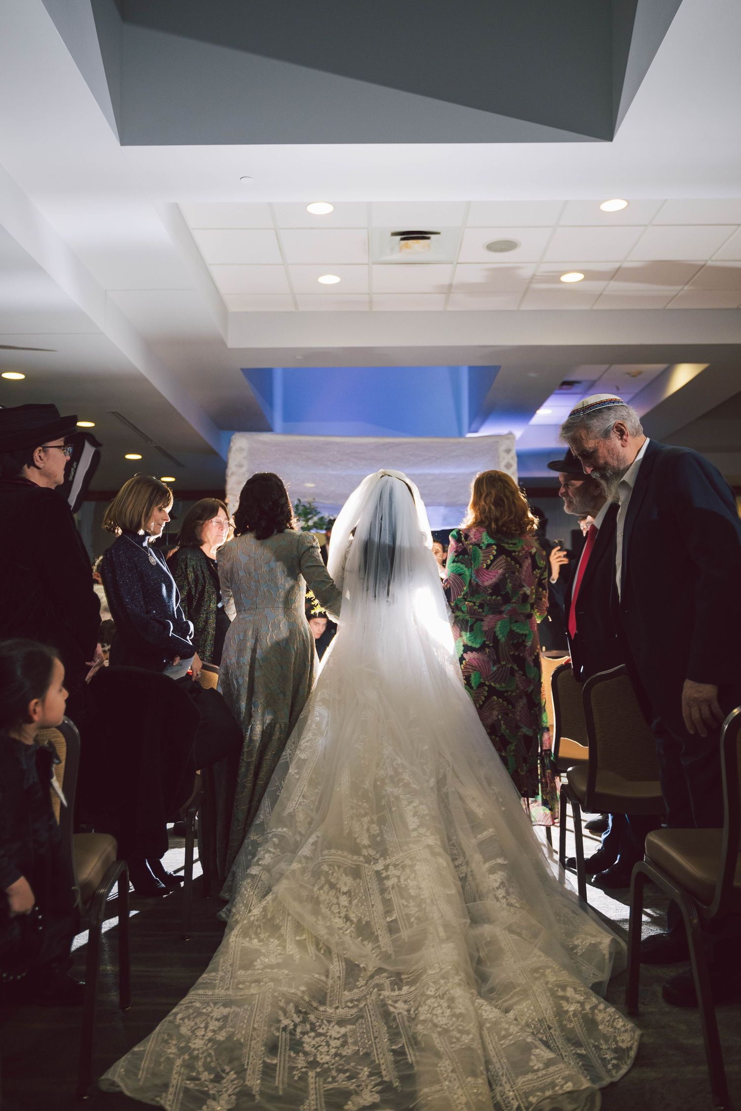 A cathedral-length wedding dress train flows down an aisle surrounded by guests.