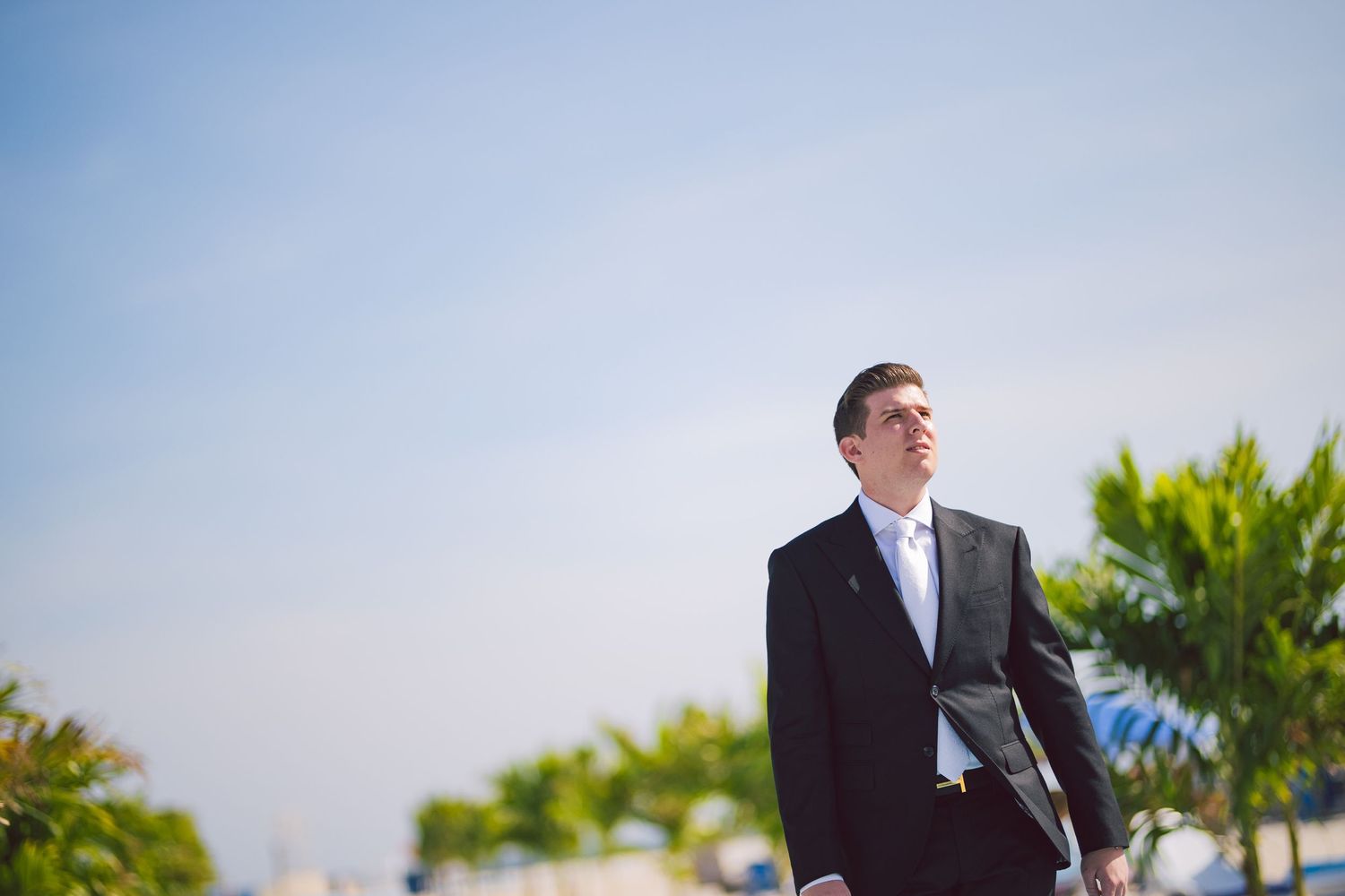 A formal outdoor wedding scene with people in suits against a bright sky and palm trees.