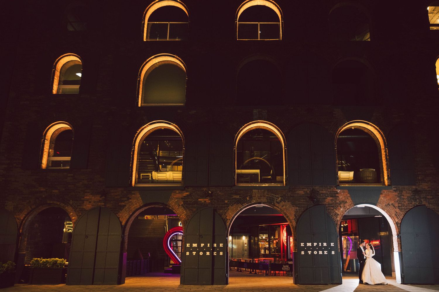 Couple shares romantic moment outside illuminated historic brick wedding venue at night.