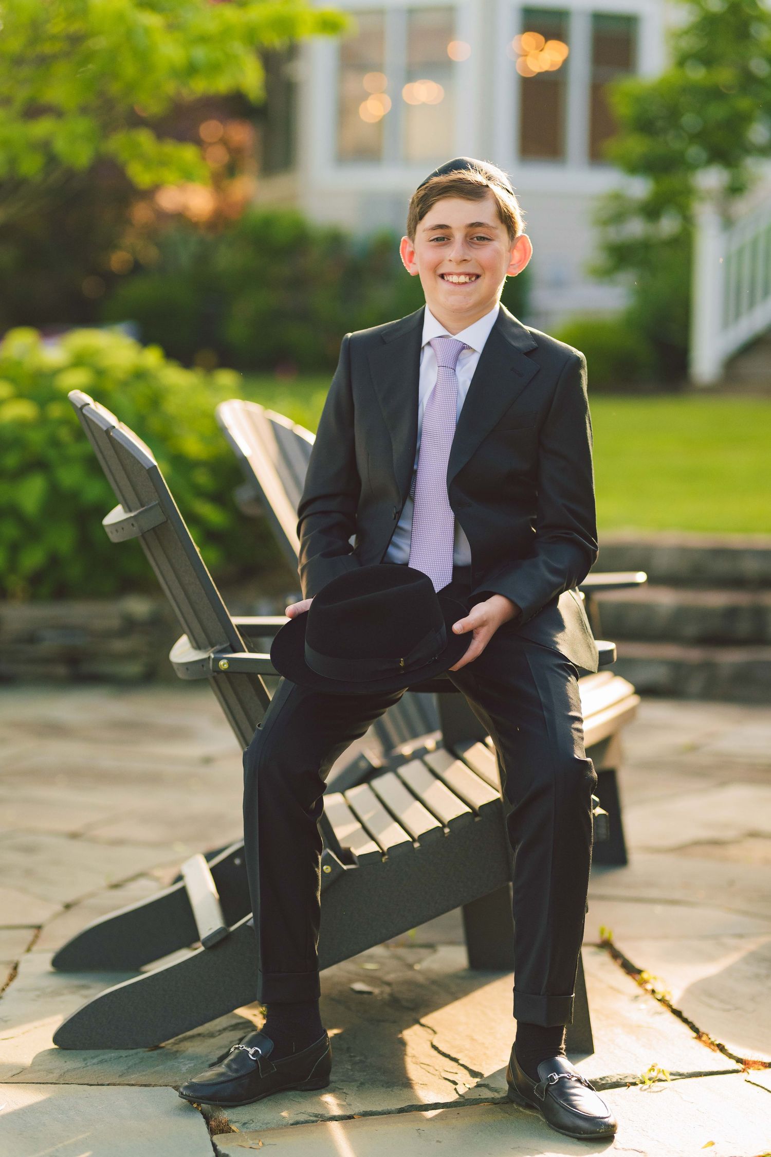 Smiling boy in dark suit and lavender tie sitting on Adirondack chair outdoors at formal event.