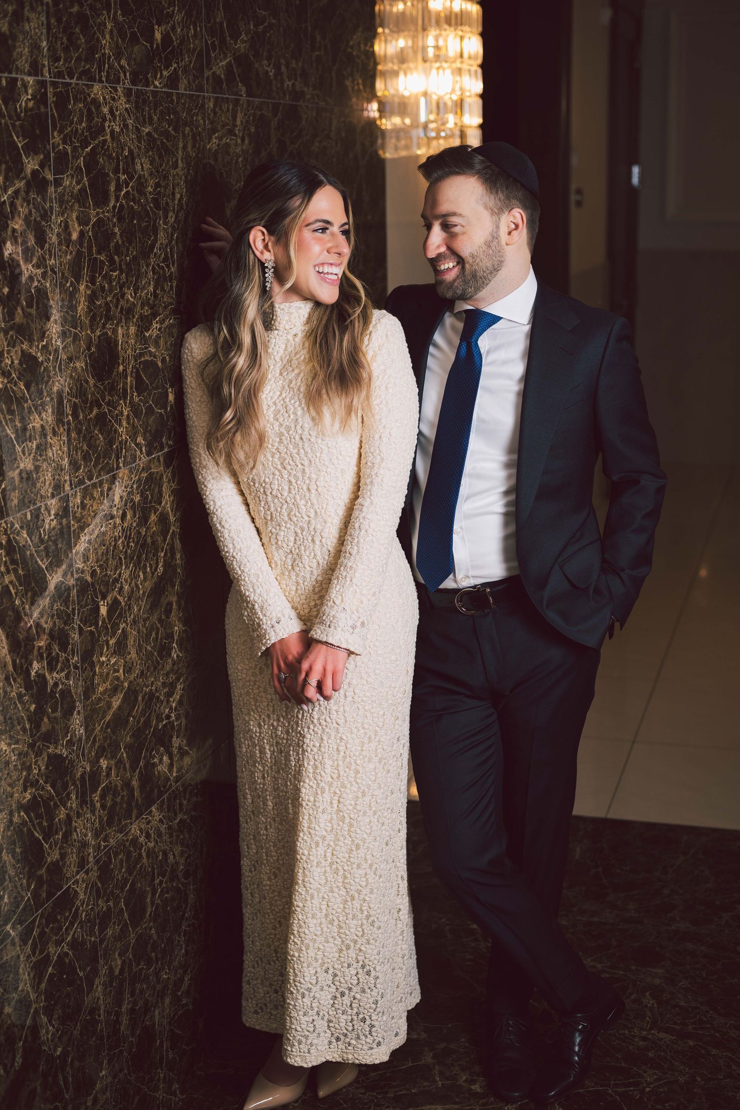 A couple shares a tender moment against a marbled wall with elegant chandeliers overhead.