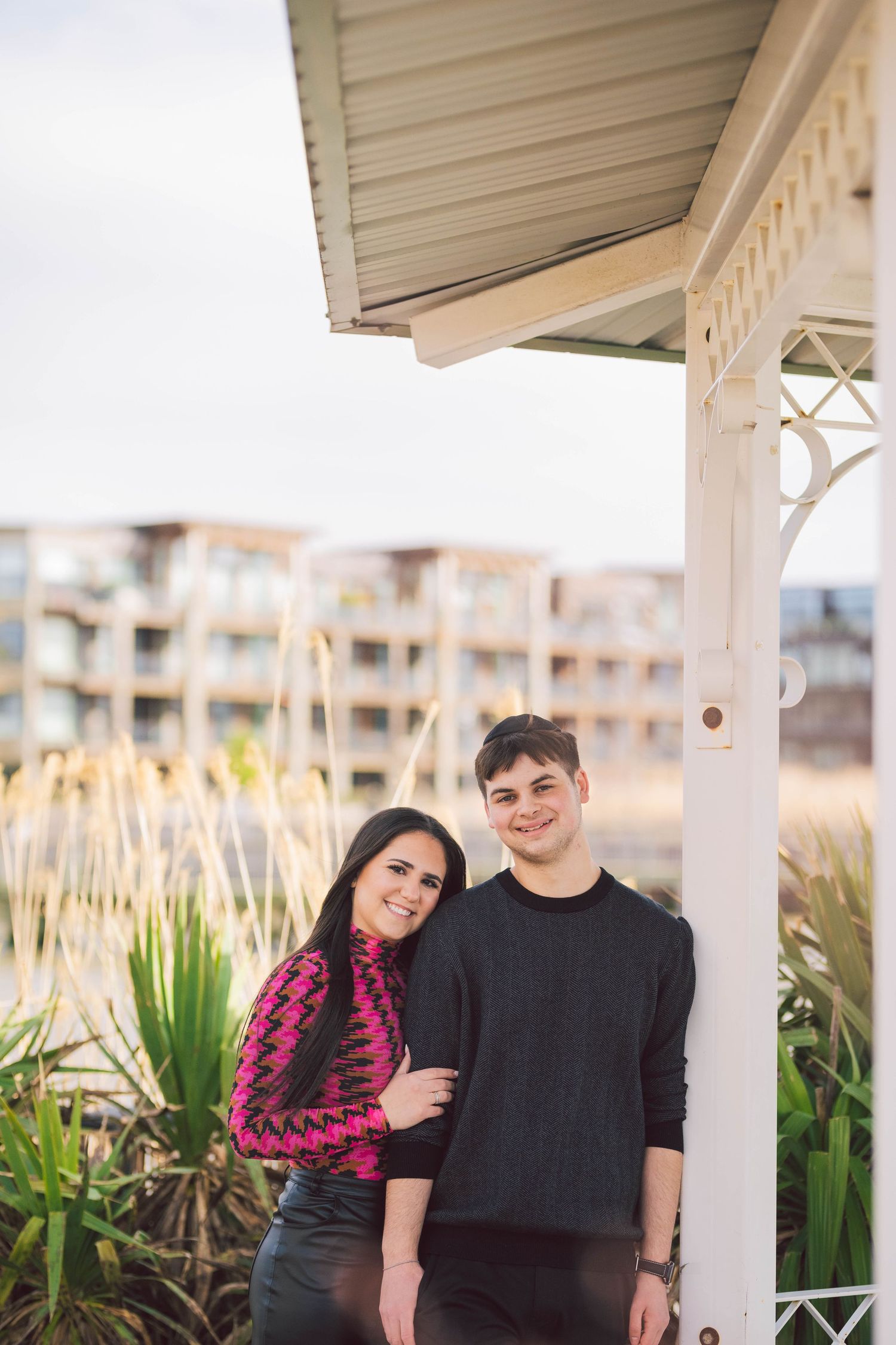 A couple poses together under a covered outdoor patio with tropical plants in the background during sunset.