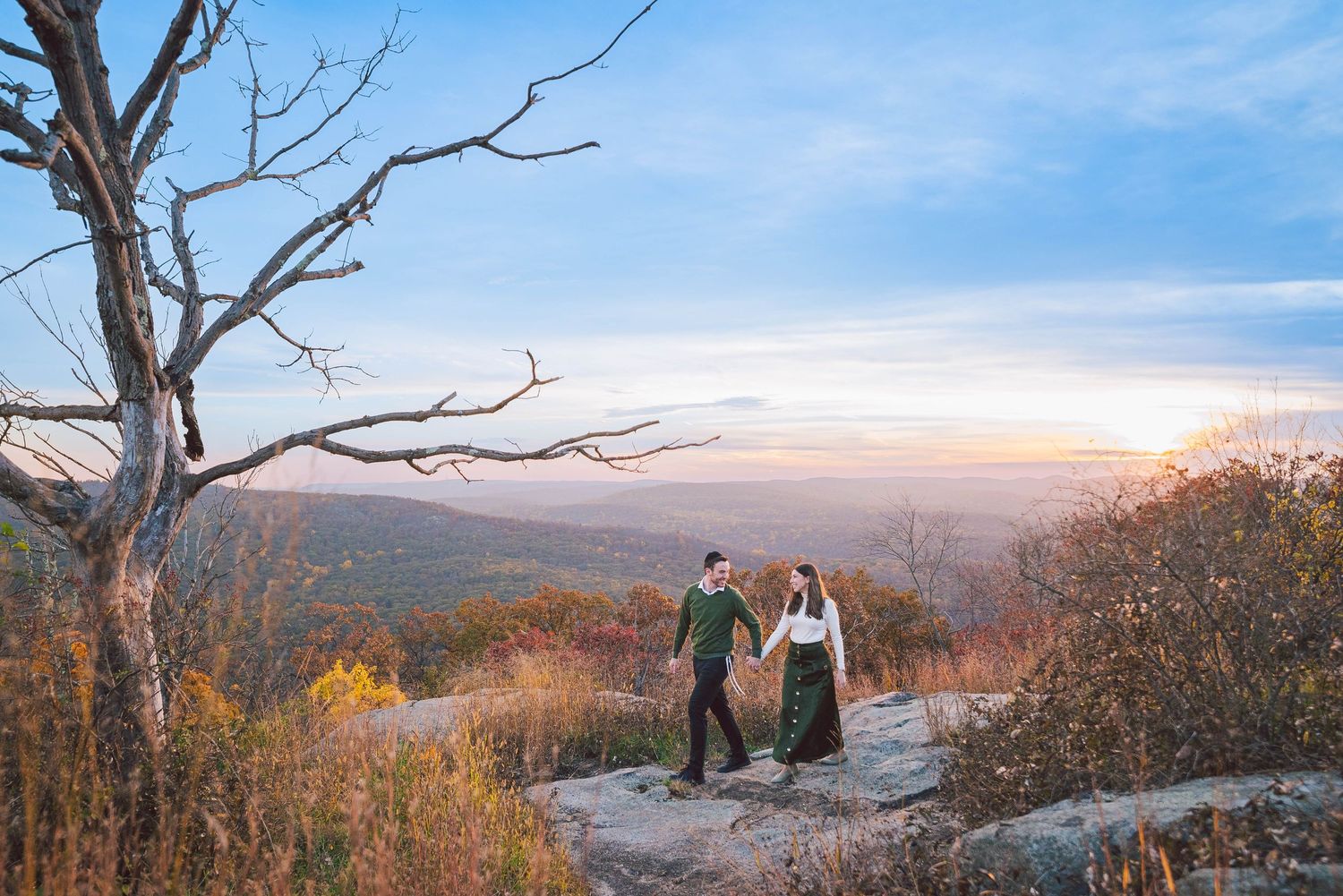 Hikers stand on rocky outcrop overlooking scenic mountain valley with autumn foliage and distant peaks at sunset.
