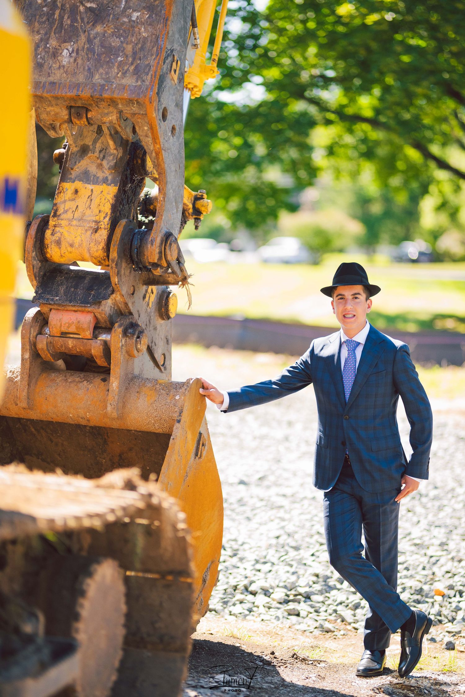 A figure in a dark suit and hat poses playfully near yellow construction equipment in an outdoor setting.