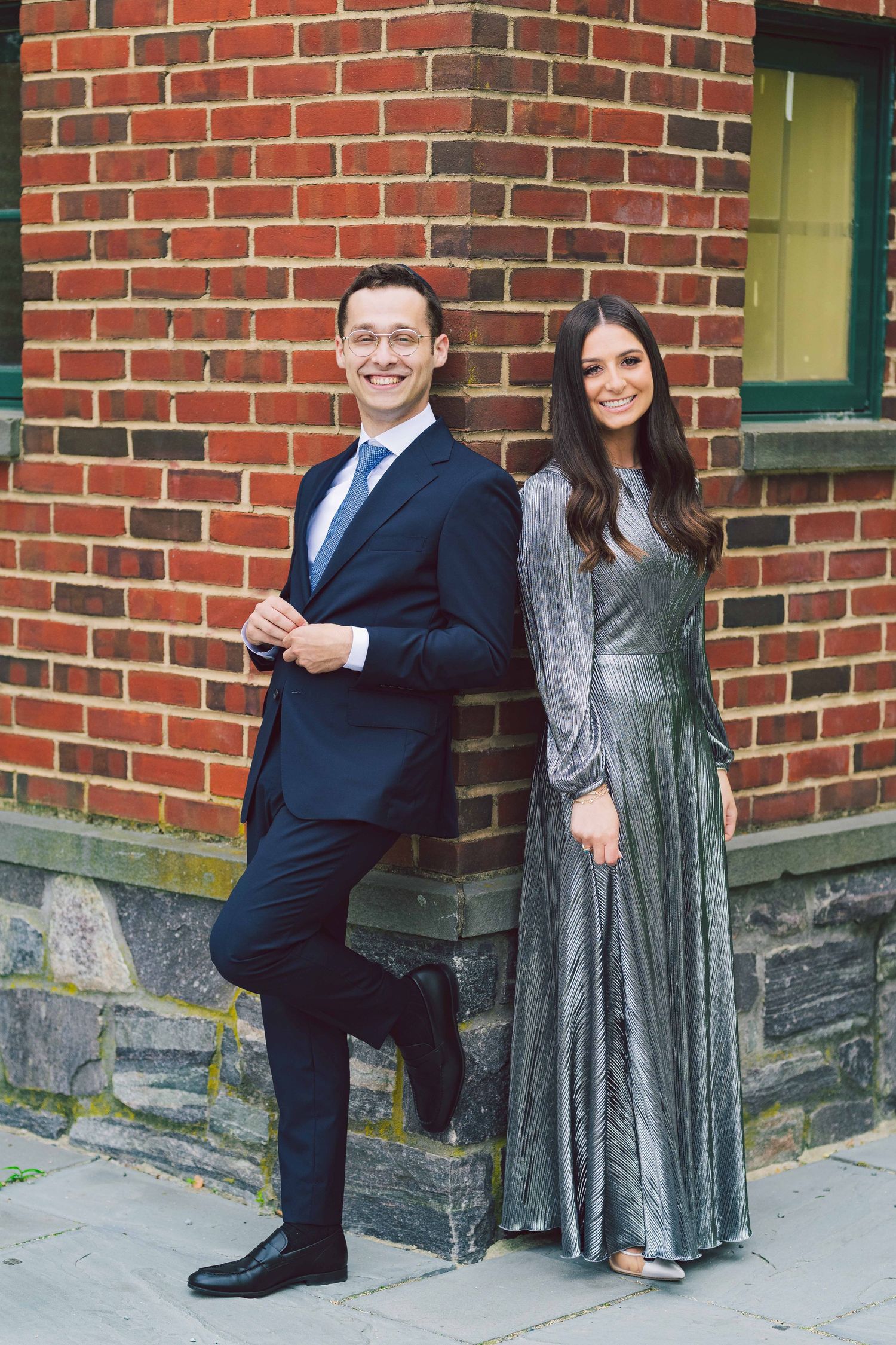 Couple poses playfully against a brick wall in formal attire.