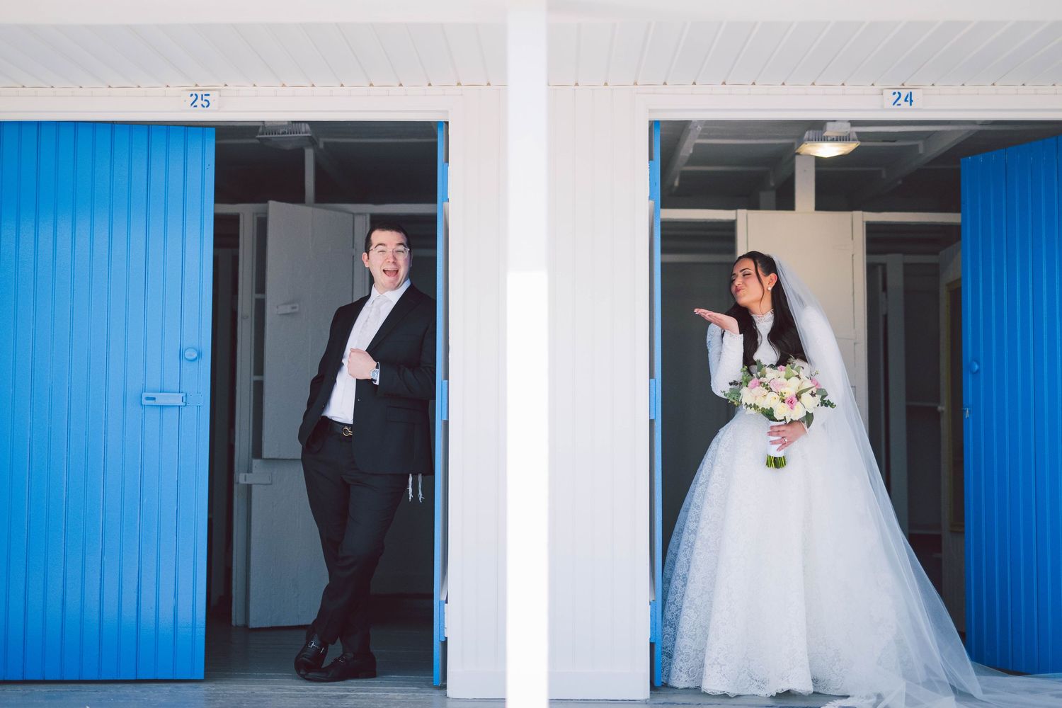 Bride in white gown and groom in black tuxedo pose separately by blue doors for wedding photos.