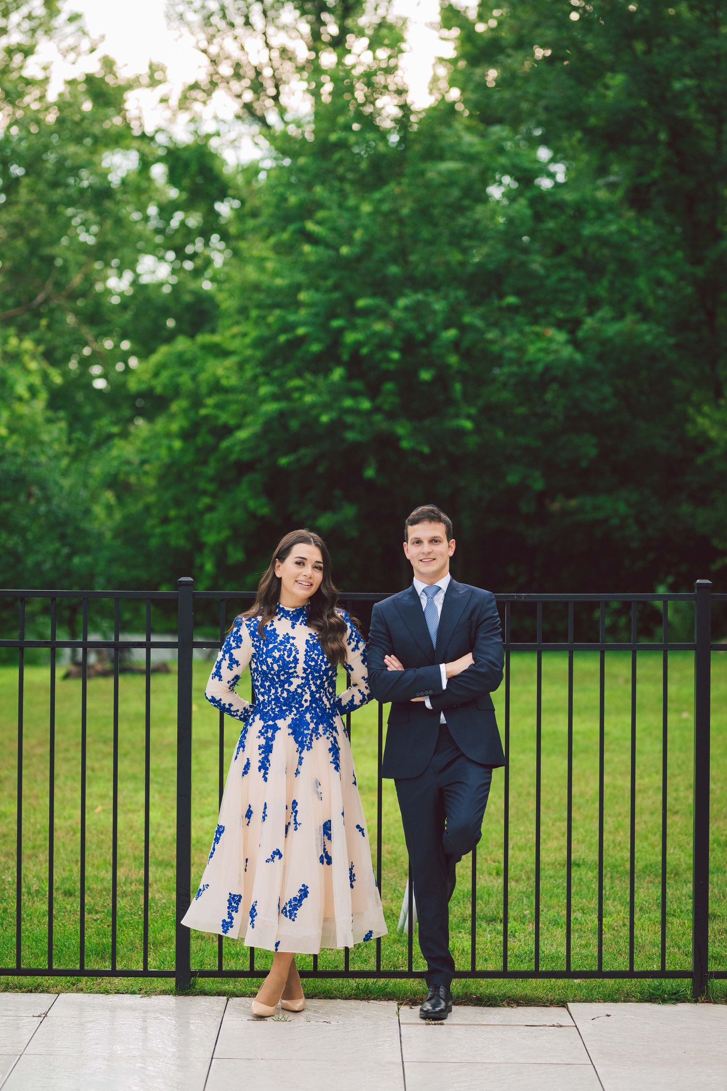 A couple poses outdoors in formal wear against a green garden backdrop with iron fencing.