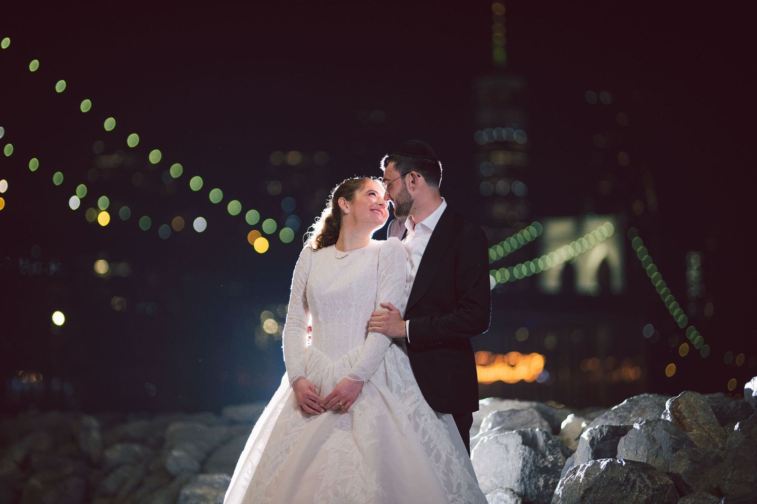 Bride and groom share a romantic moment at night with city lights and bokeh in the background.