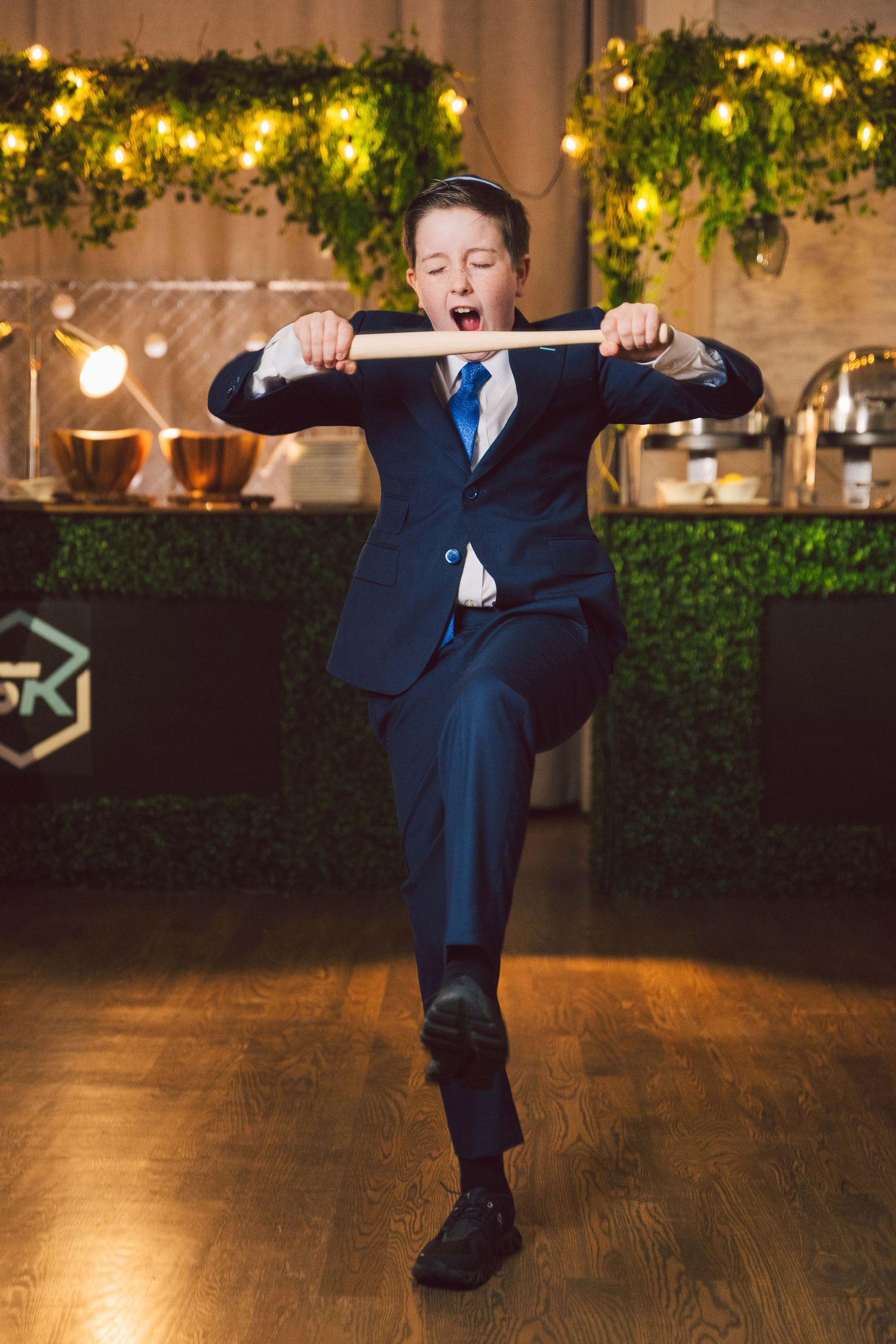 Young guest dancing energetically at an event with decorative greenery and lights in background.