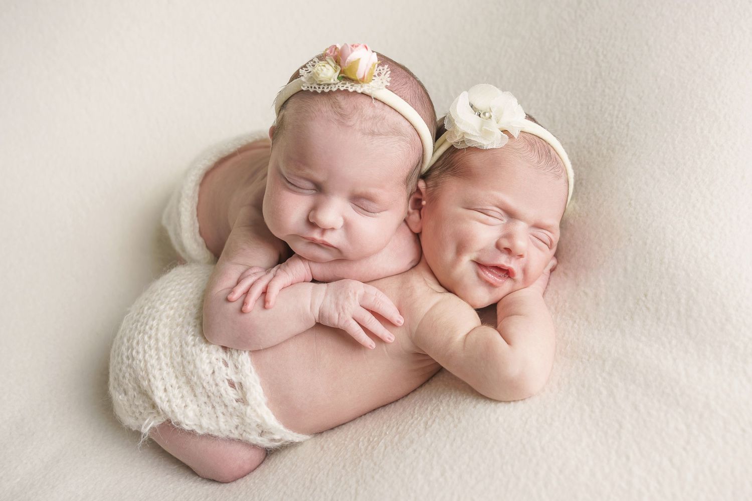 Twin newborns sleeping together wearing floral headbands on cream backdrop.