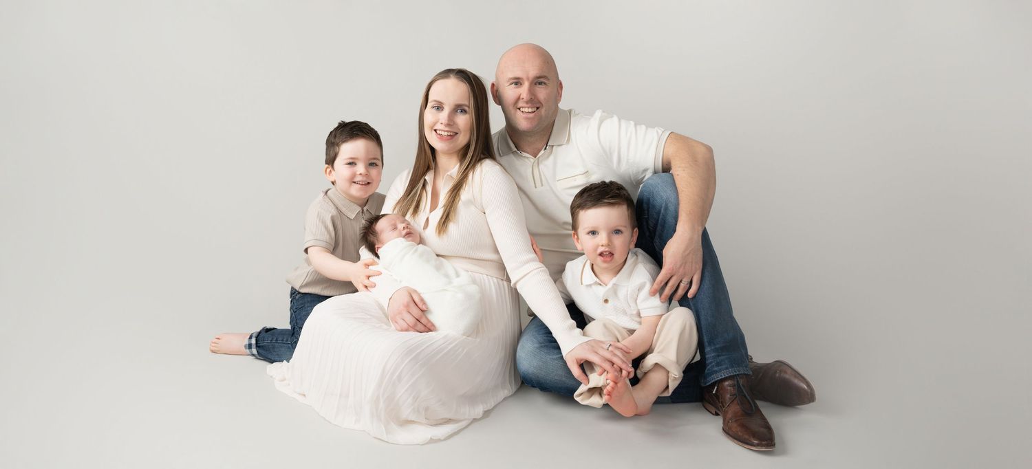 Family studio portrait with pregnant mother in white dress against light background.