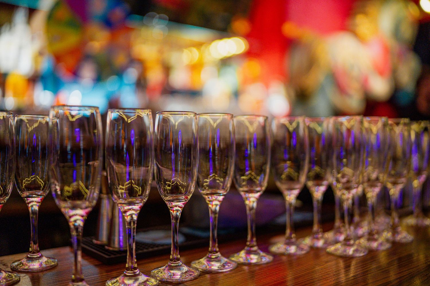Row of empty wine glasses lined up on a bar with colorful blurred party lights in the background.