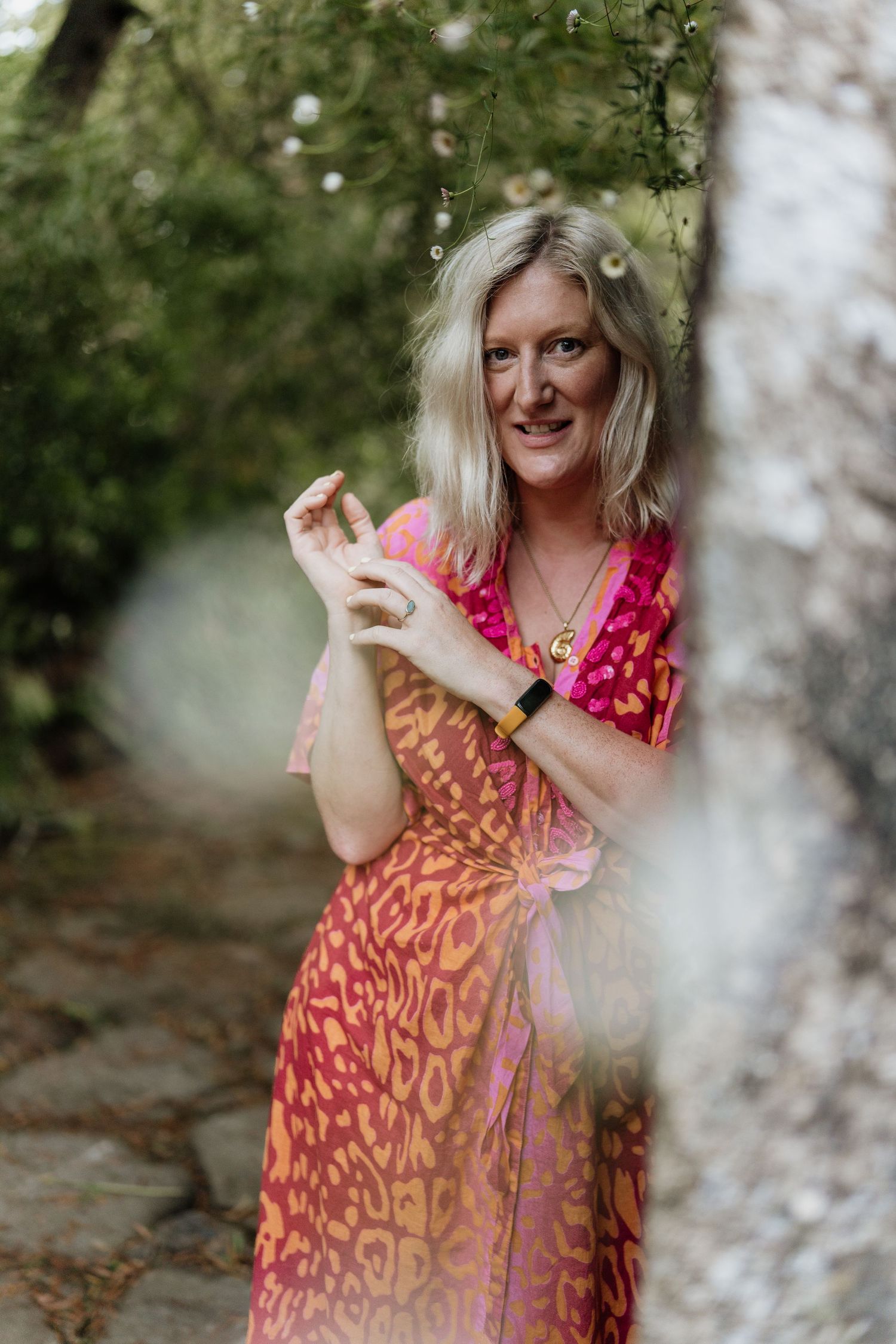 Person in pink and orange patterned dress poses playfully in an outdoor garden setting.
