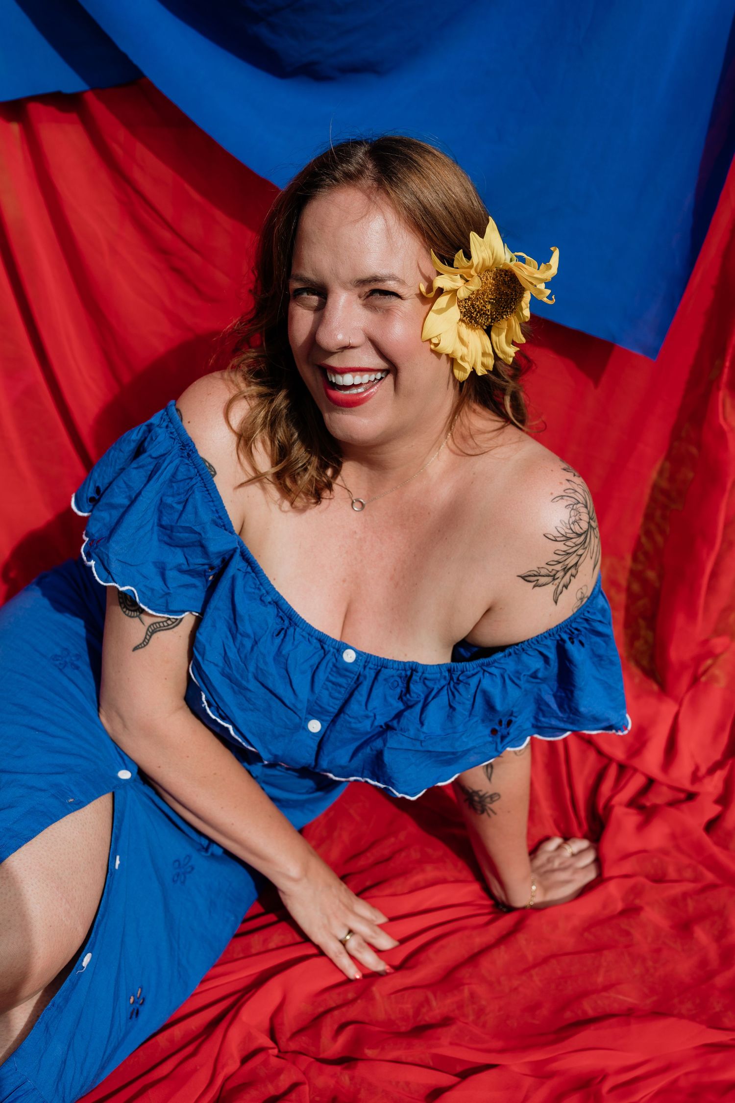 Portrait against red and blue backdrop featuring vibrant blue off-shoulder dress and yellow sunflower hair accessory.