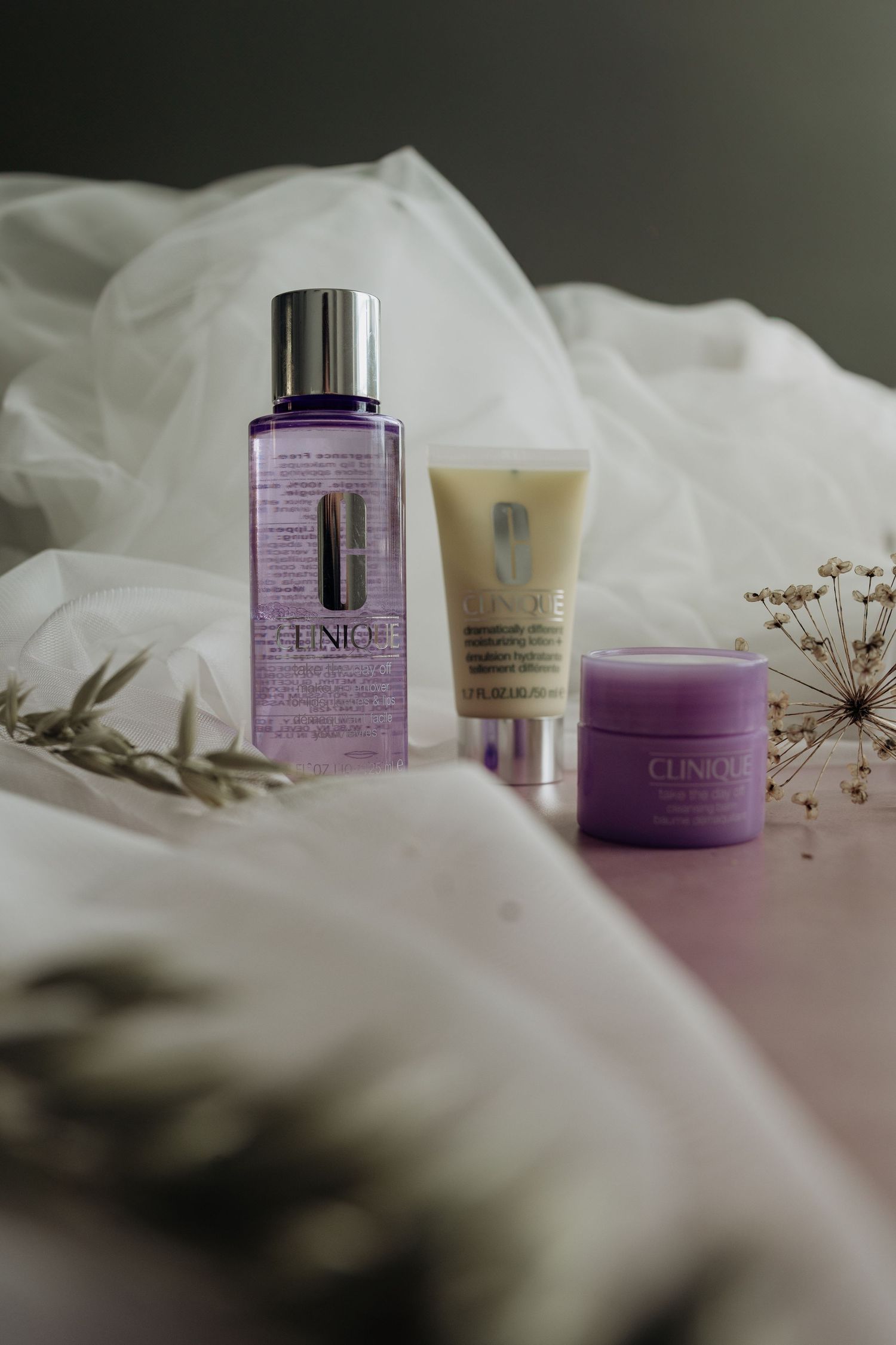 Purple skincare cosmetic jars and bottles arranged with white fabric and dried floral elements in a soft, dreamy composition.