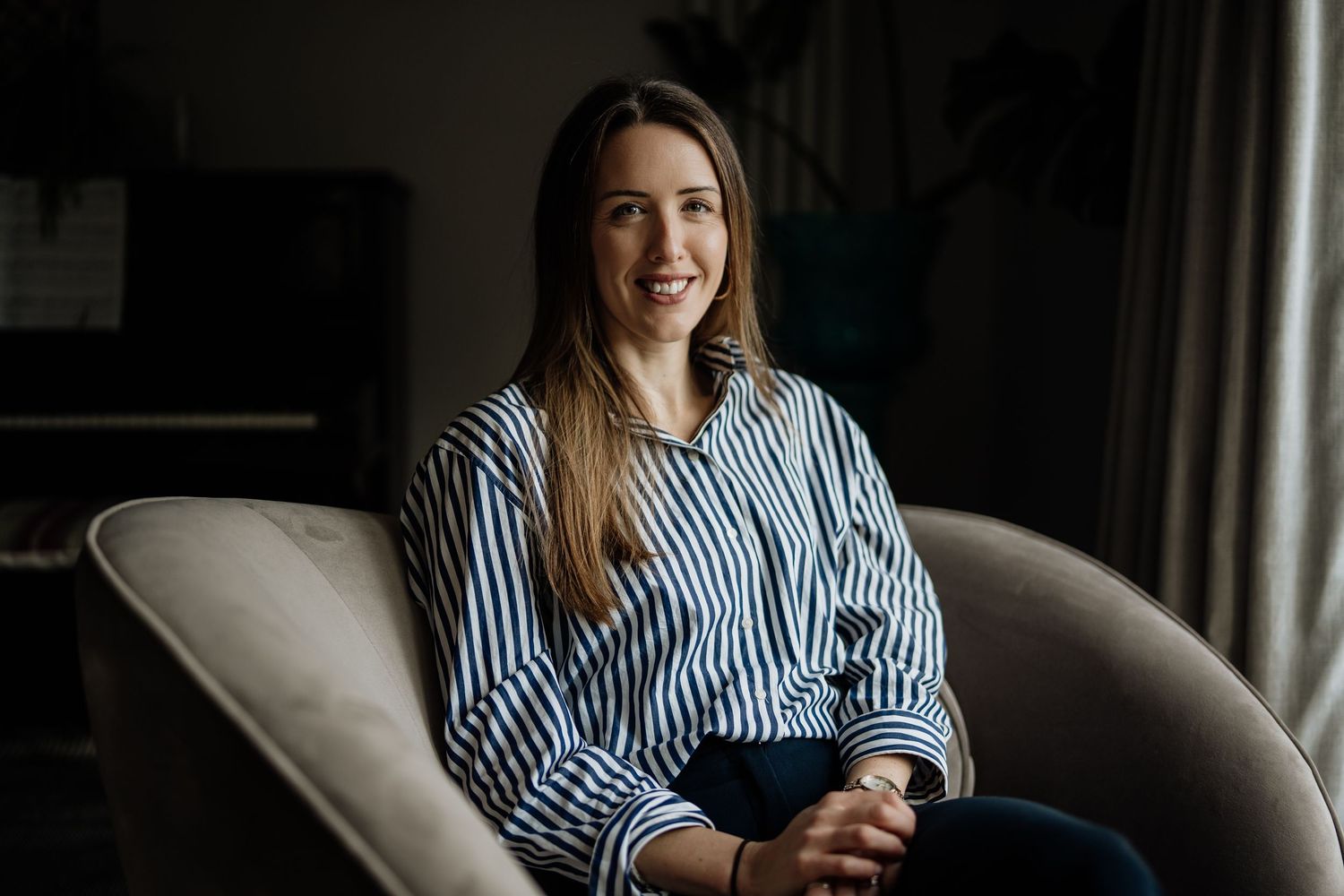 A smiling person in a blue and white striped shirt sits comfortably on a gray couch in a dark room.