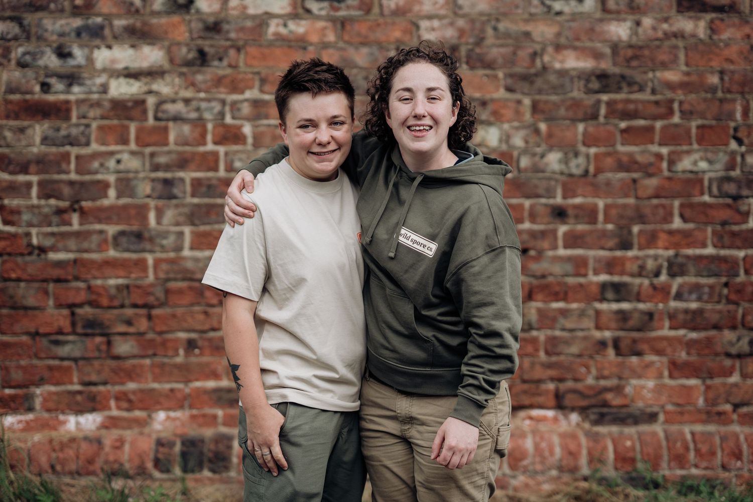 Two young people smile while standing together against a rustic brick wall background in casual clothing.