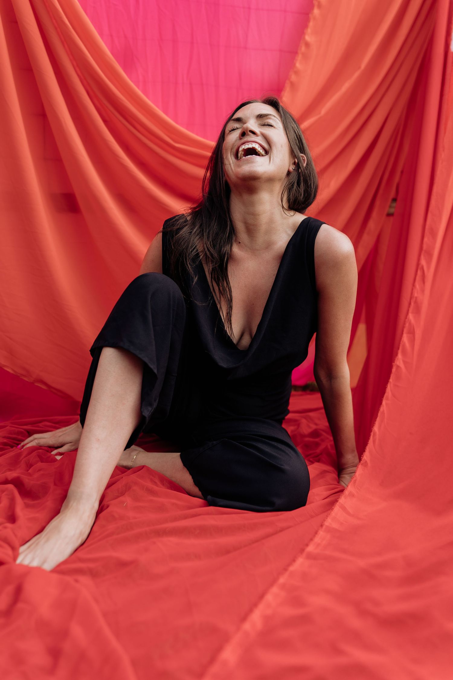 A joyful moment captured against a vibrant red backdrop as someone in a black dress laughs freely while sitting on the floor.