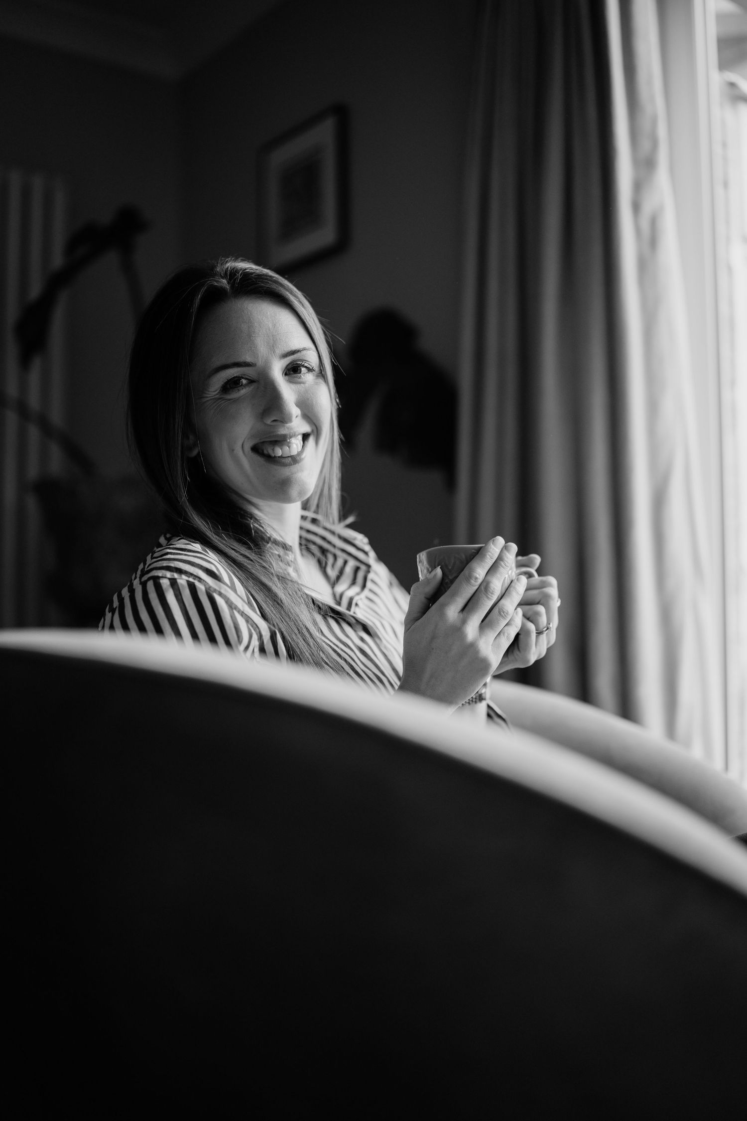 A candid black and white photo shows a smiling figure holding a coffee mug near a window with natural lighting.