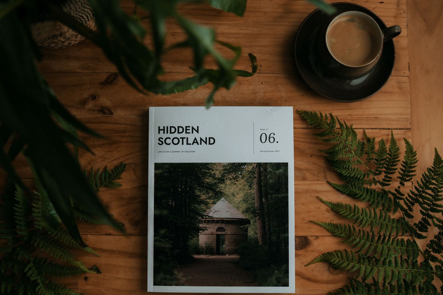 Hidden Scotland magazine issue 06 lies on a wooden surface with fern leaves and coffee cup in moody lighting.