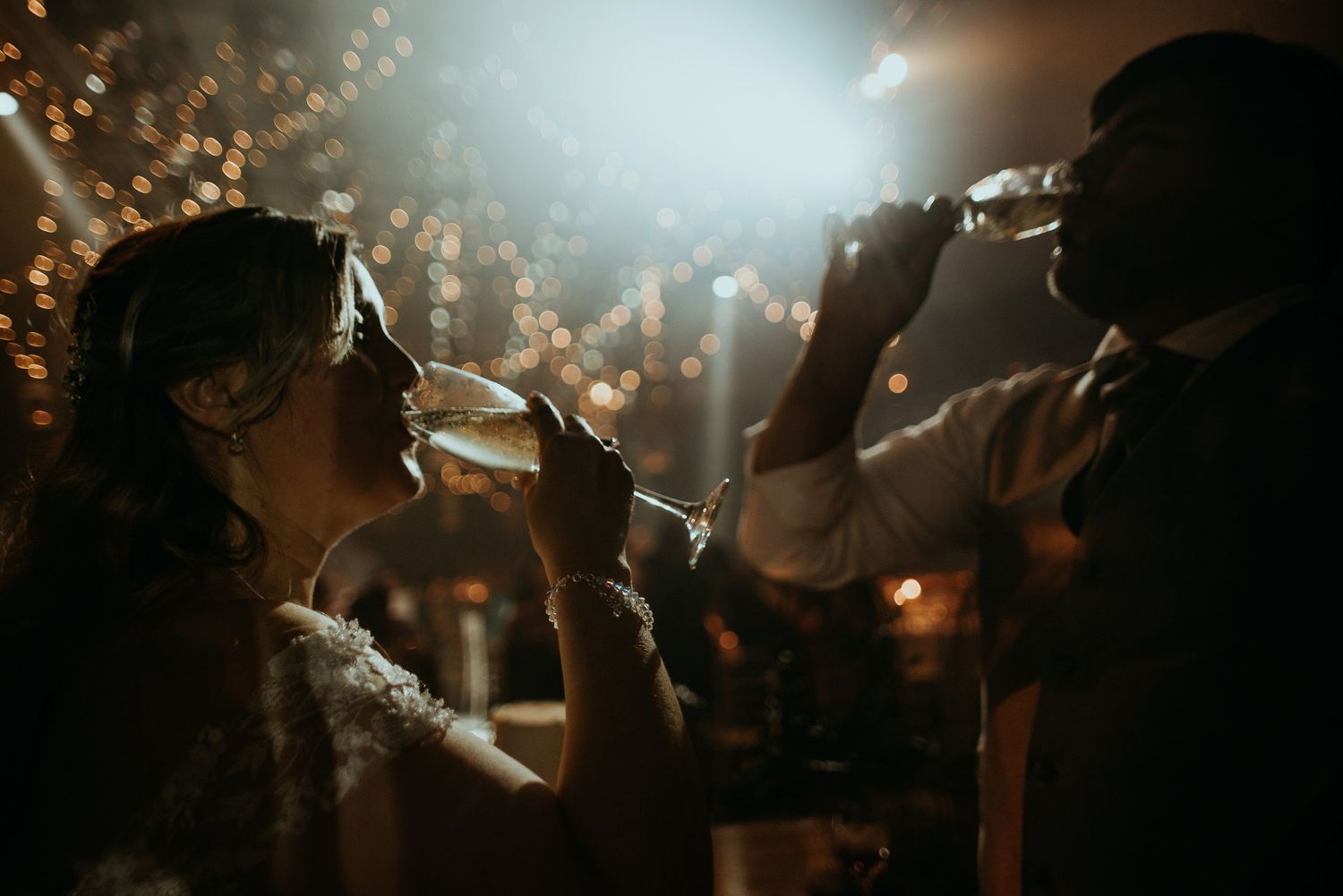 Bride and groom sharing a romantic champagne toast at their wedding reception with bokeh lights.