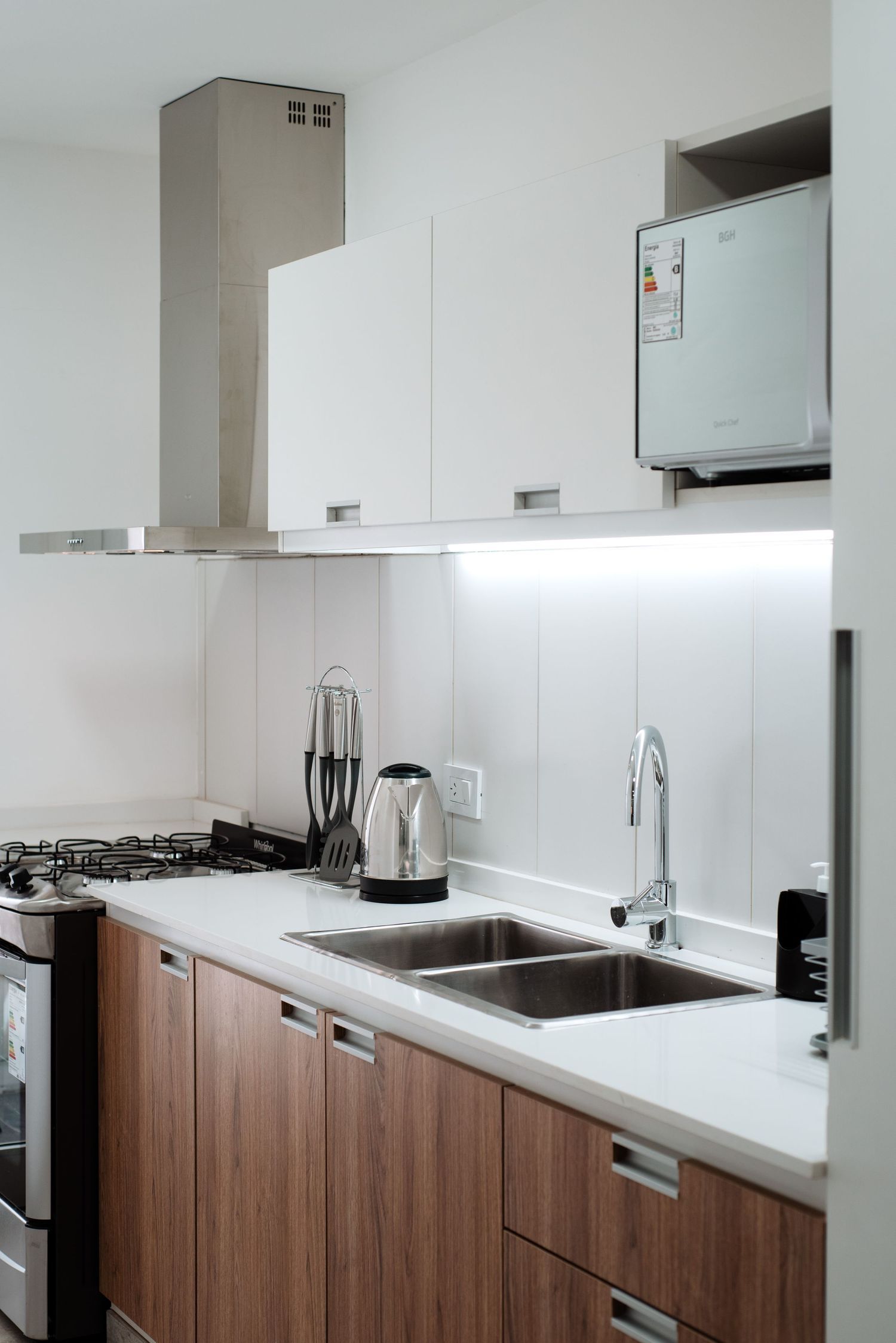 Modern kitchen with stainless steel sink, wood cabinets, gas stove, range hood, and white countertops.