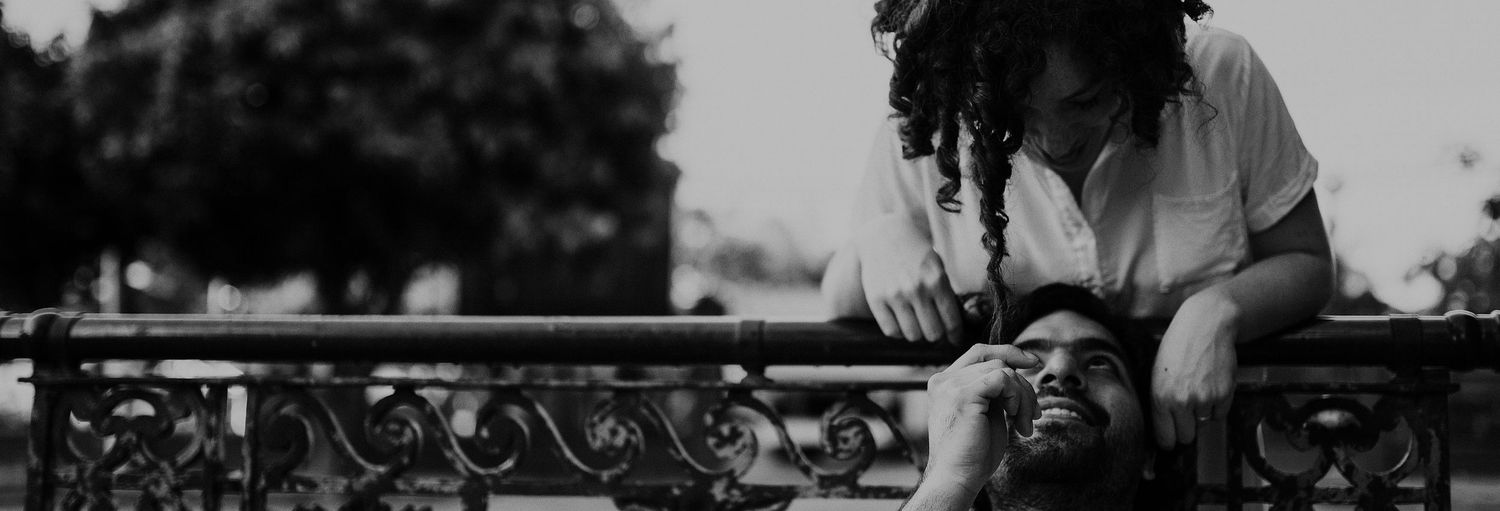 A couple shares a tender moment leaning over an ornate iron railing in a black and white outdoor photo.