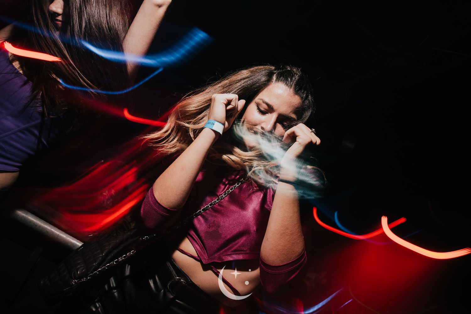 Young woman dancing at a nightclub with colorful neon light trails in a dark party atmosphere.