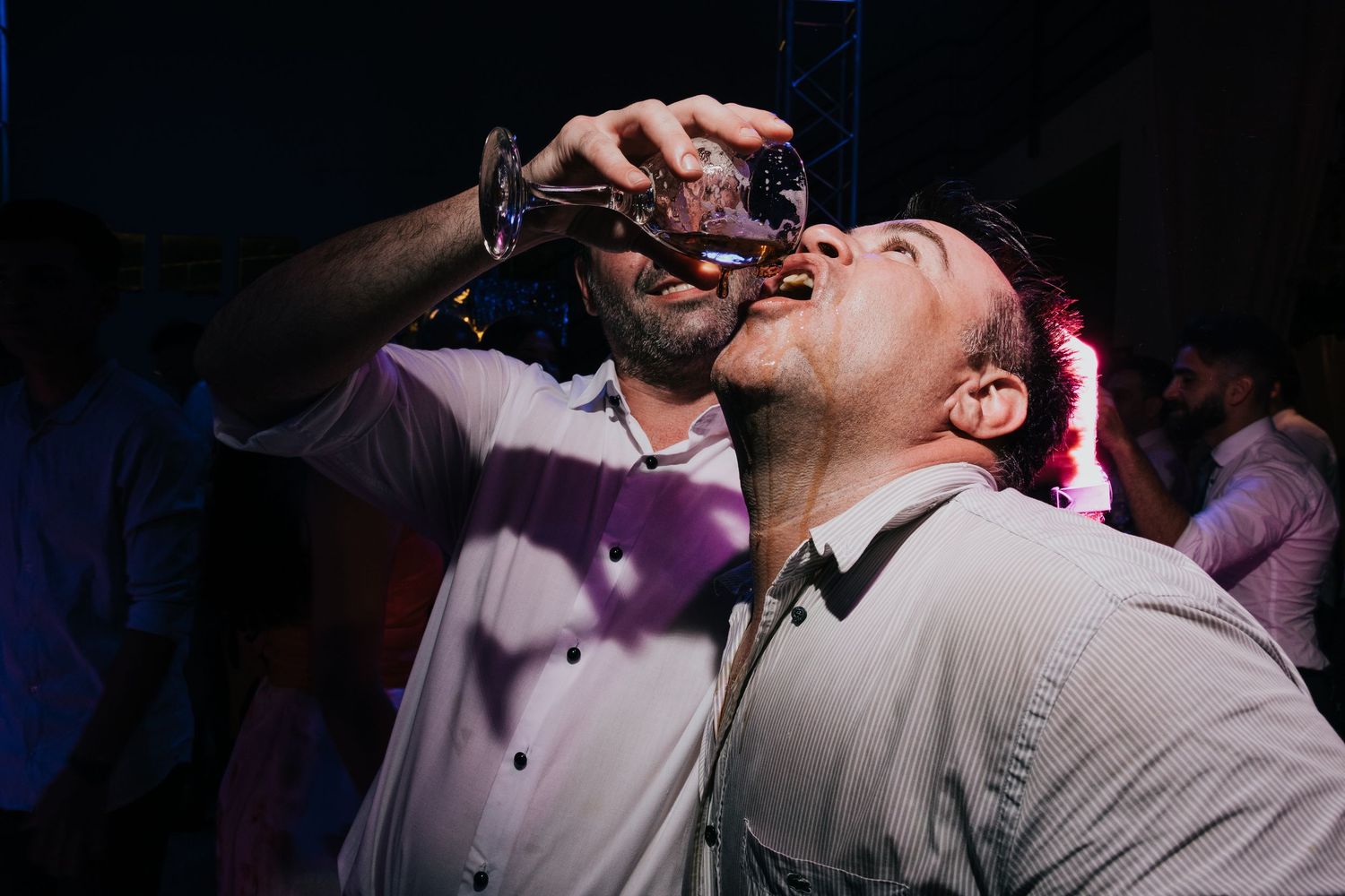 Two men drinking shots at a lively party event under dramatic lighting.