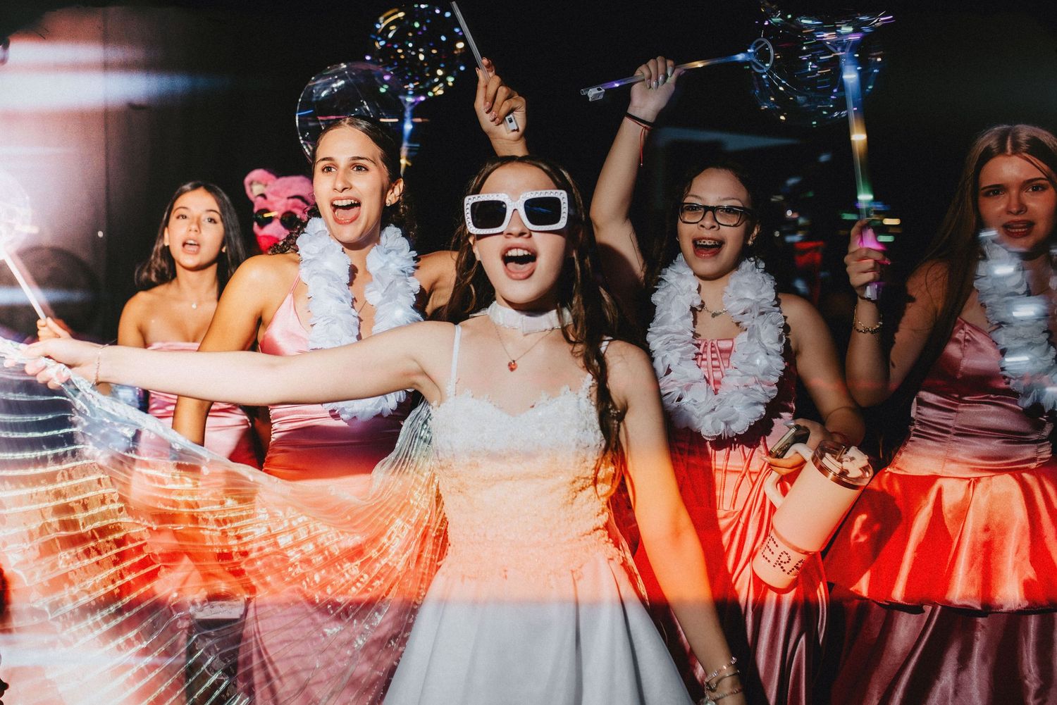 Joyful bride and friends dancing at wedding reception, holding light-up wands and wearing feather boas.