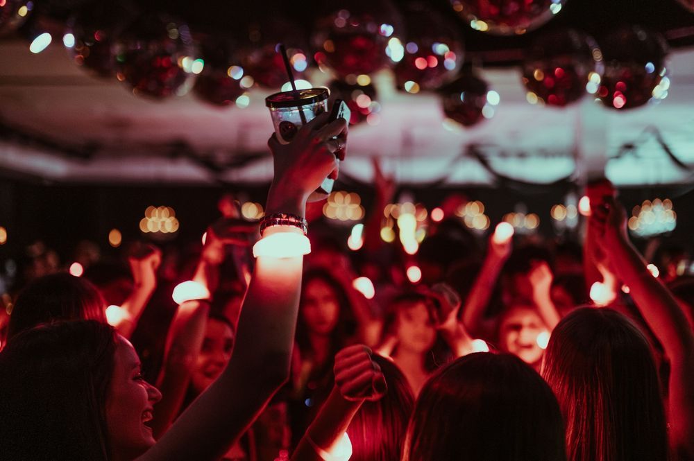 Crowd of people dancing at a festive party with red lights, disco balls, and a hand holding a drink raised up.