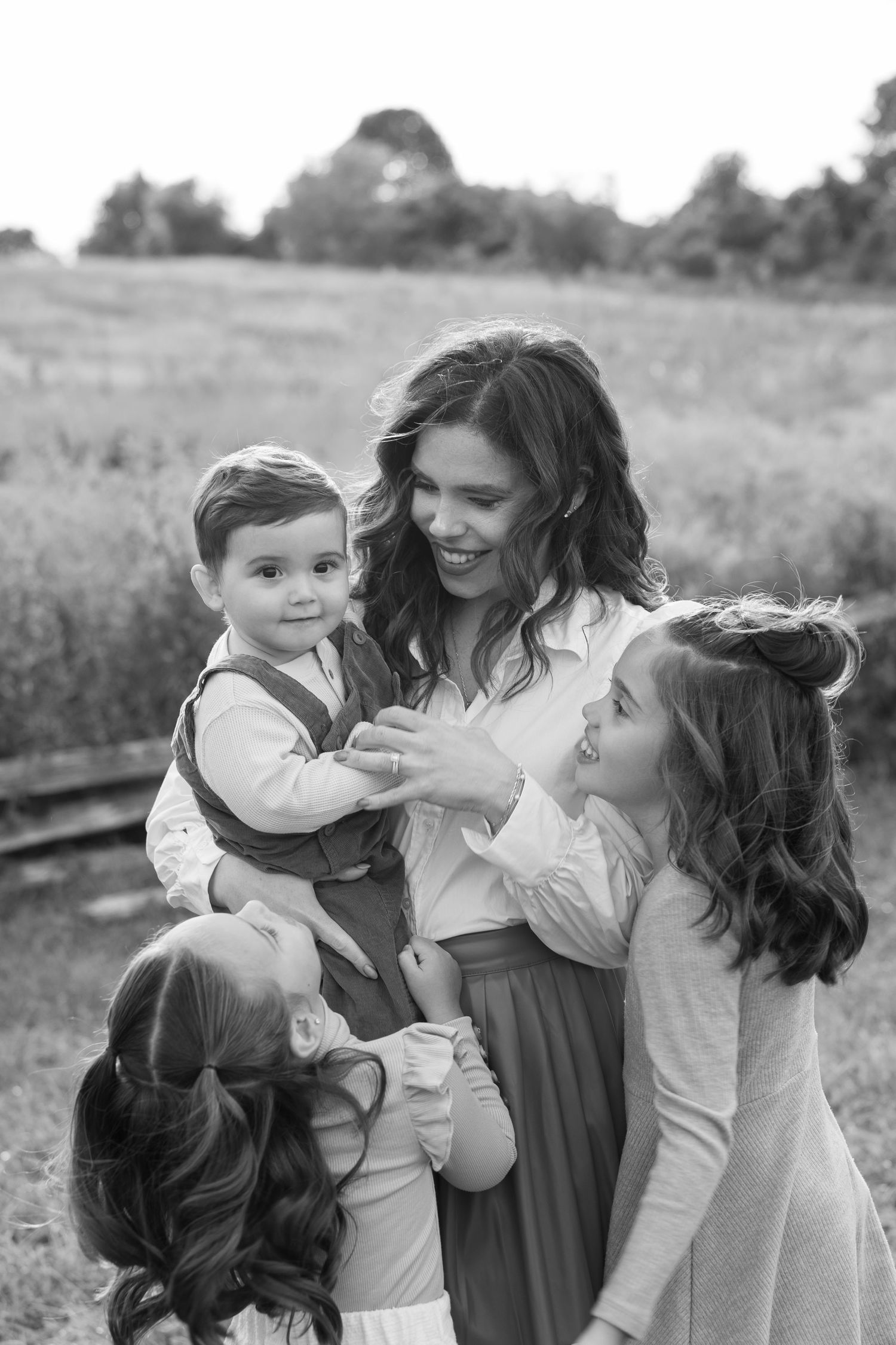 Black and white photo of a smiling mother holding her toddler son while two daughters embrace her in a field.