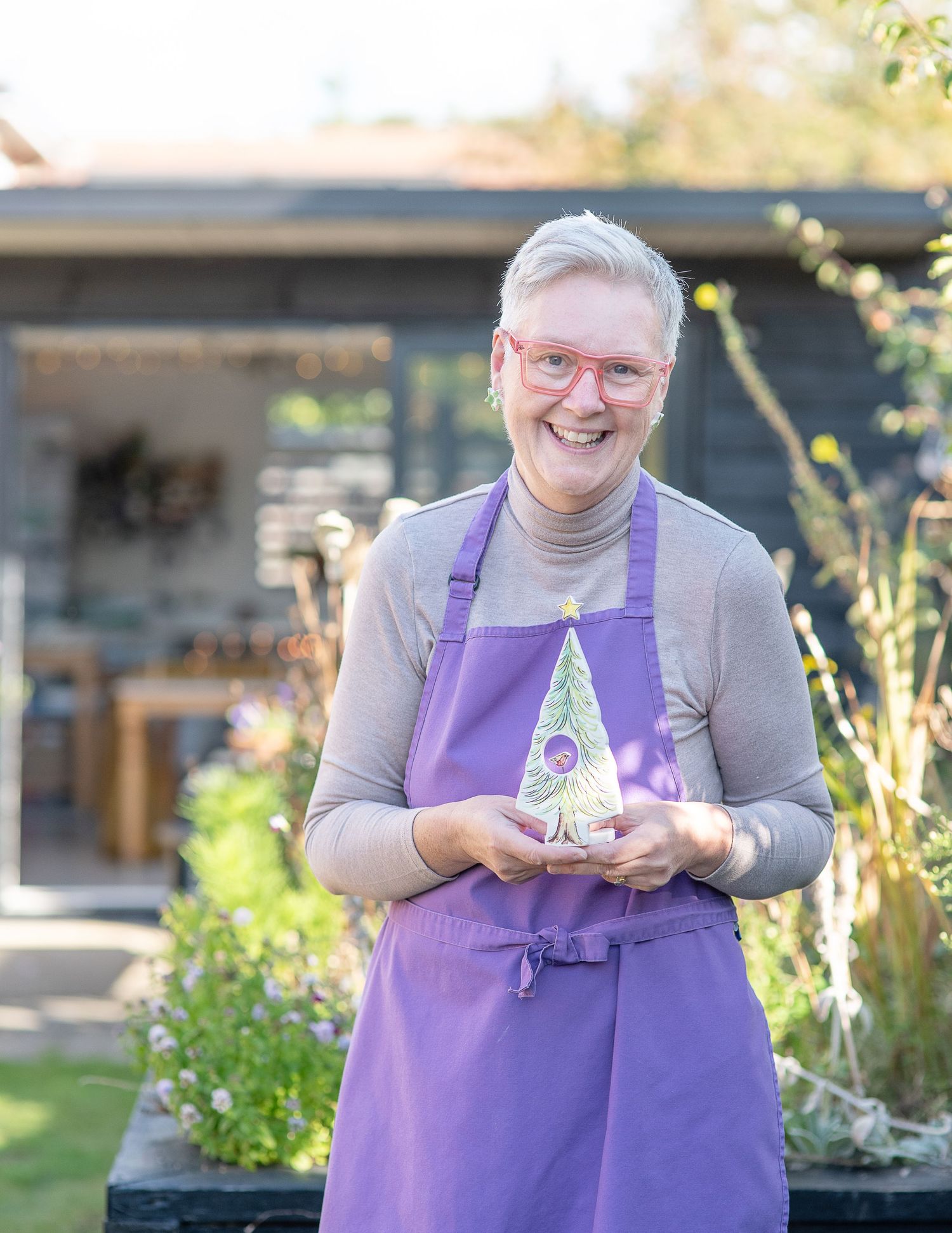 A smiling woman in a purple apron holds a decorative award in a sunny garden setting.