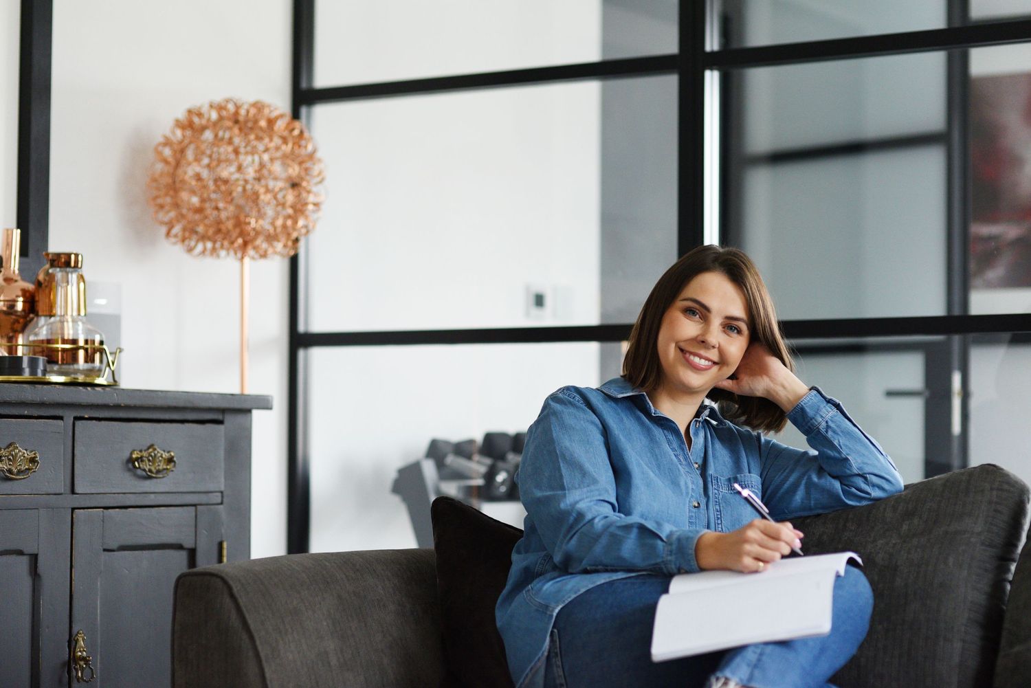 Professional in blue denim shirt reviews documents at modern office desk with large windows.