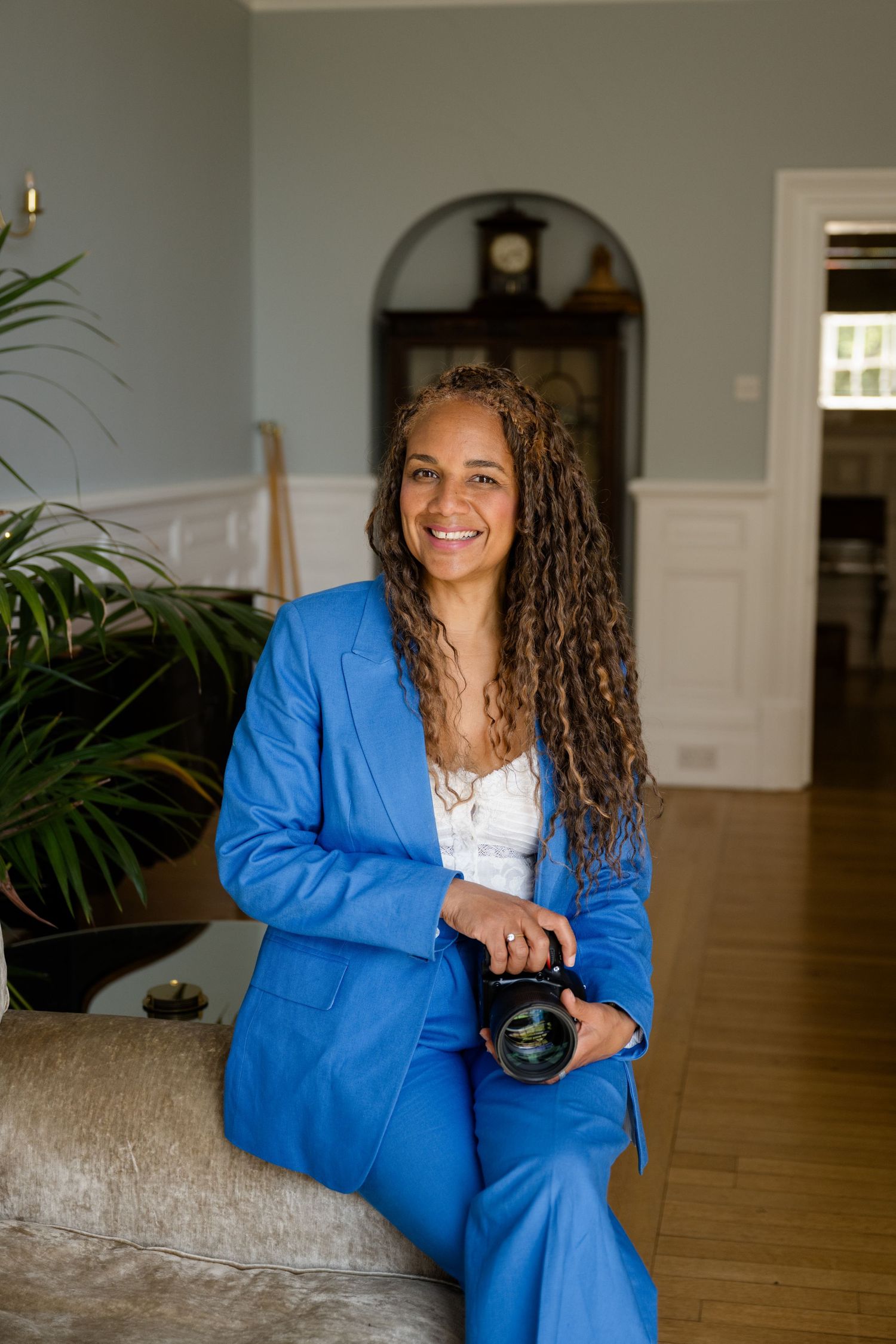 Woman in blue suit holding camera, smiling in elegant home interior with hardwood floors and arched doorway.