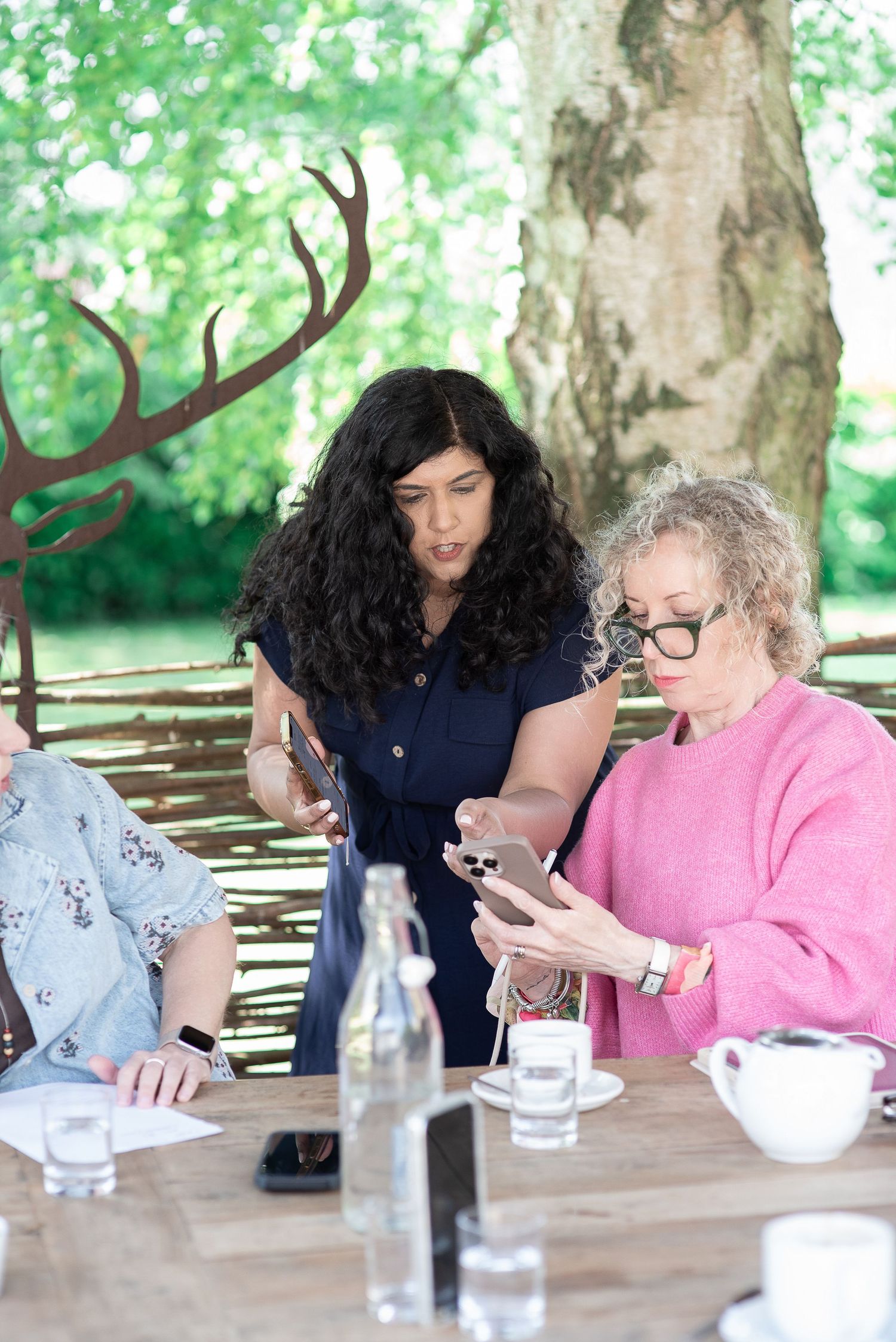 Two women at outdoor table, one with dark curly hair helping older woman in pink sweater use a smartphone.