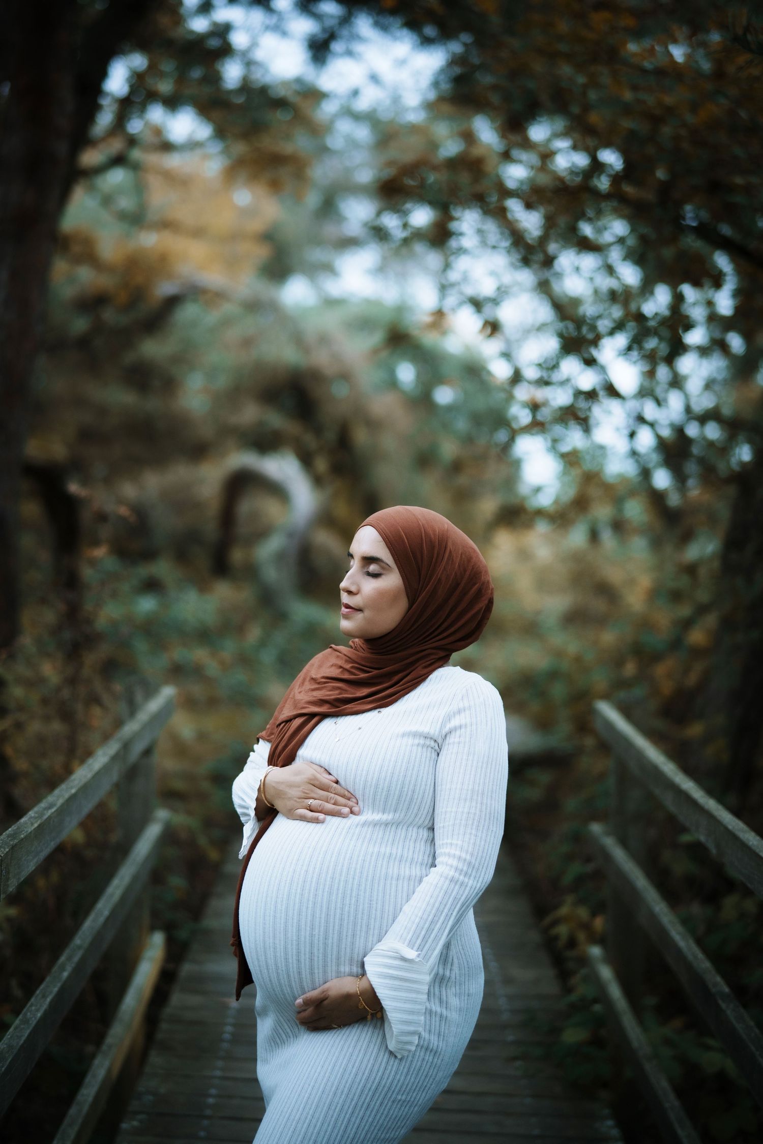 A pregnant woman in white dress and brown hijab poses thoughtfully on a wooden bridge in nature.