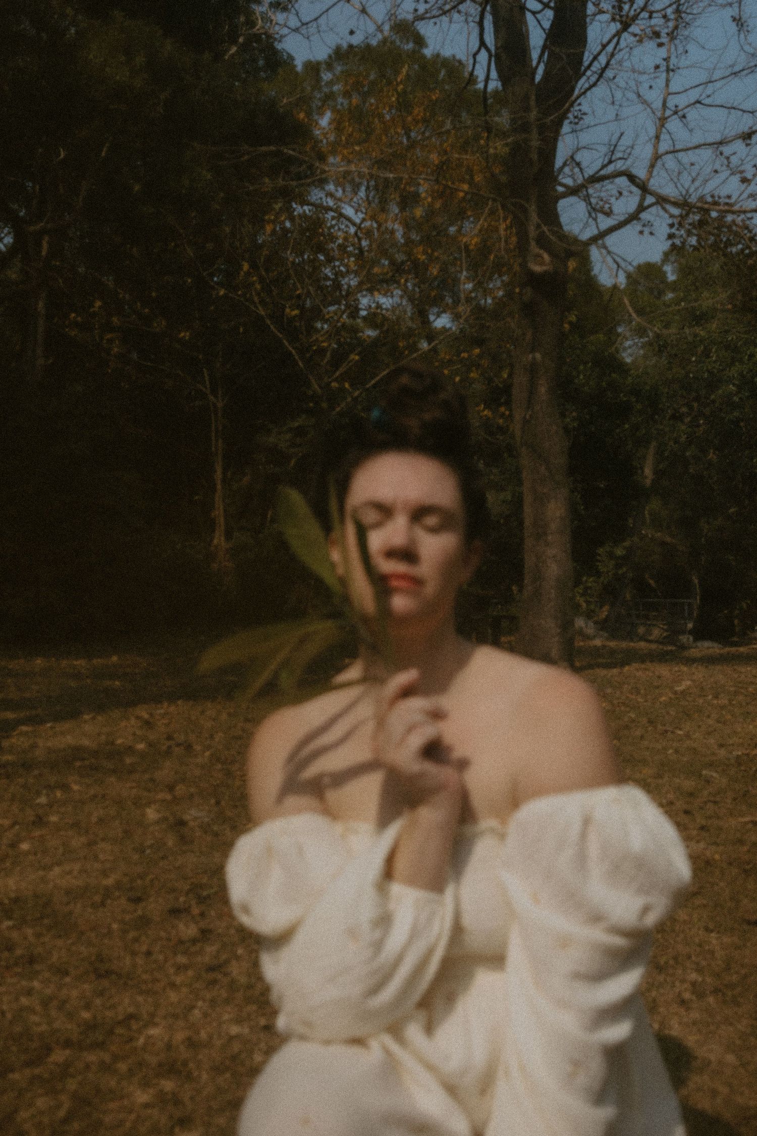 A woman in a white off-shoulder dress holds a leaf with eyes closed, surrounded by trees in golden light.