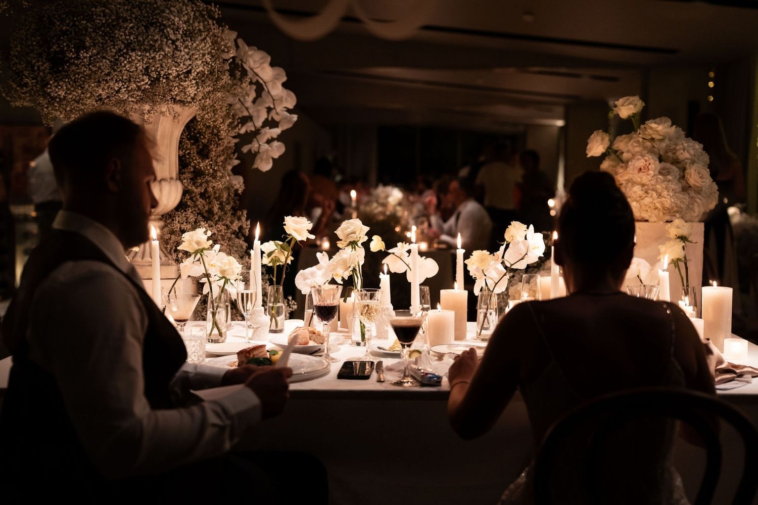 Wedding reception tables decorated with white floral arrangements and glowing candles in a dimly lit venue. - JCclick Wedding photo & Film -