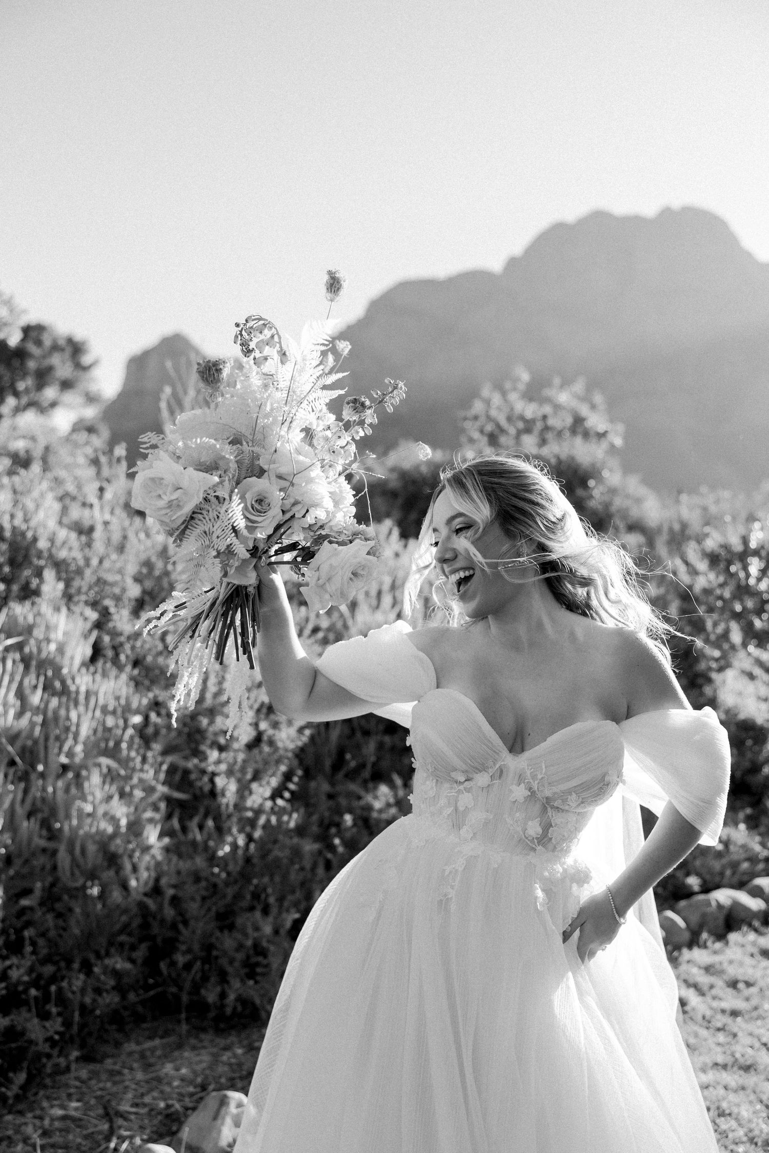 Black and white photo of bride holding bouquet against mountain landscape. - JCclick Wedding photo & Film - Best Cape Town Photographer.