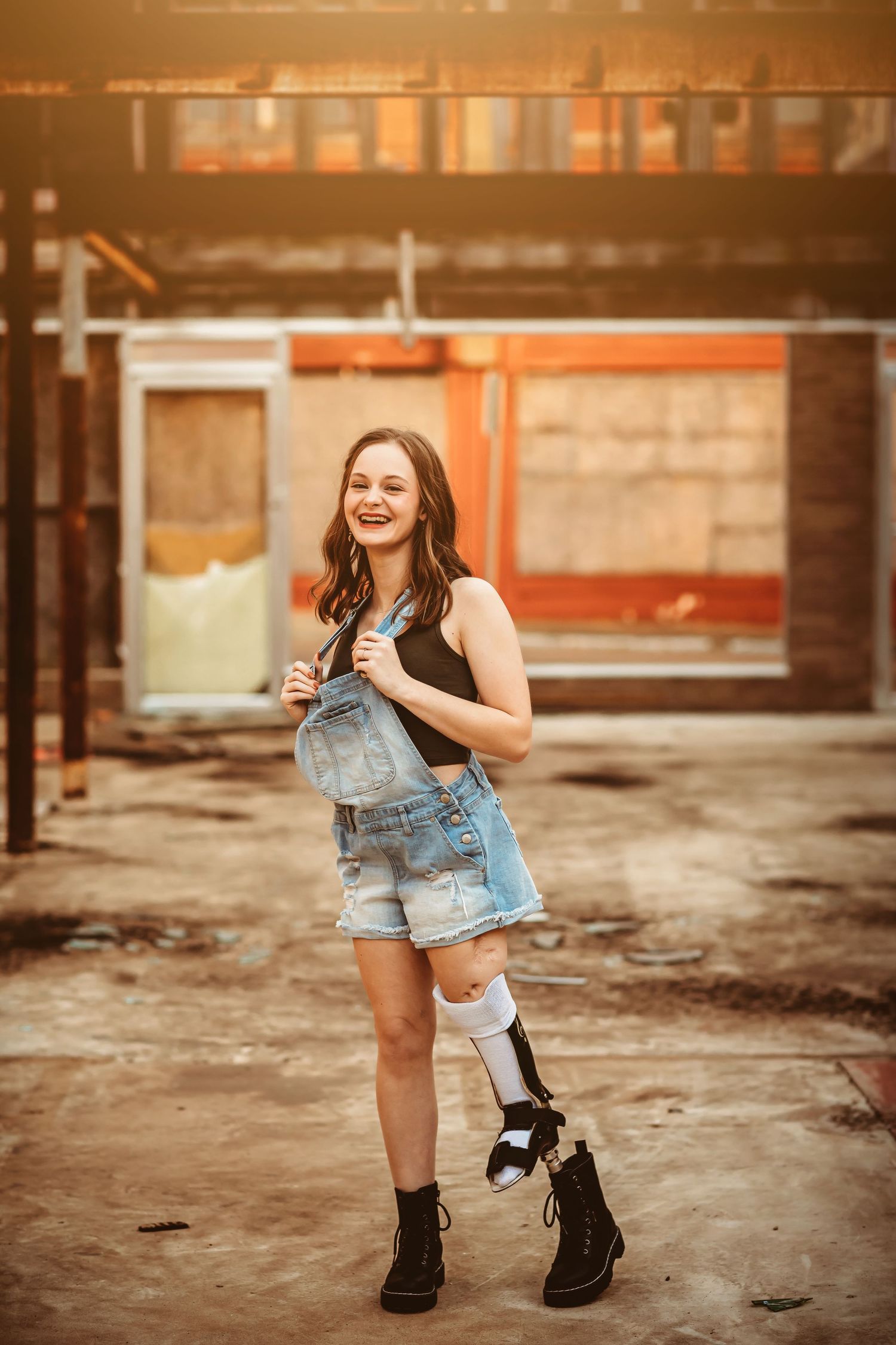 Smiling young woman with a prosthetic leg wearing denim overalls and boots, posing confidently outdoors.